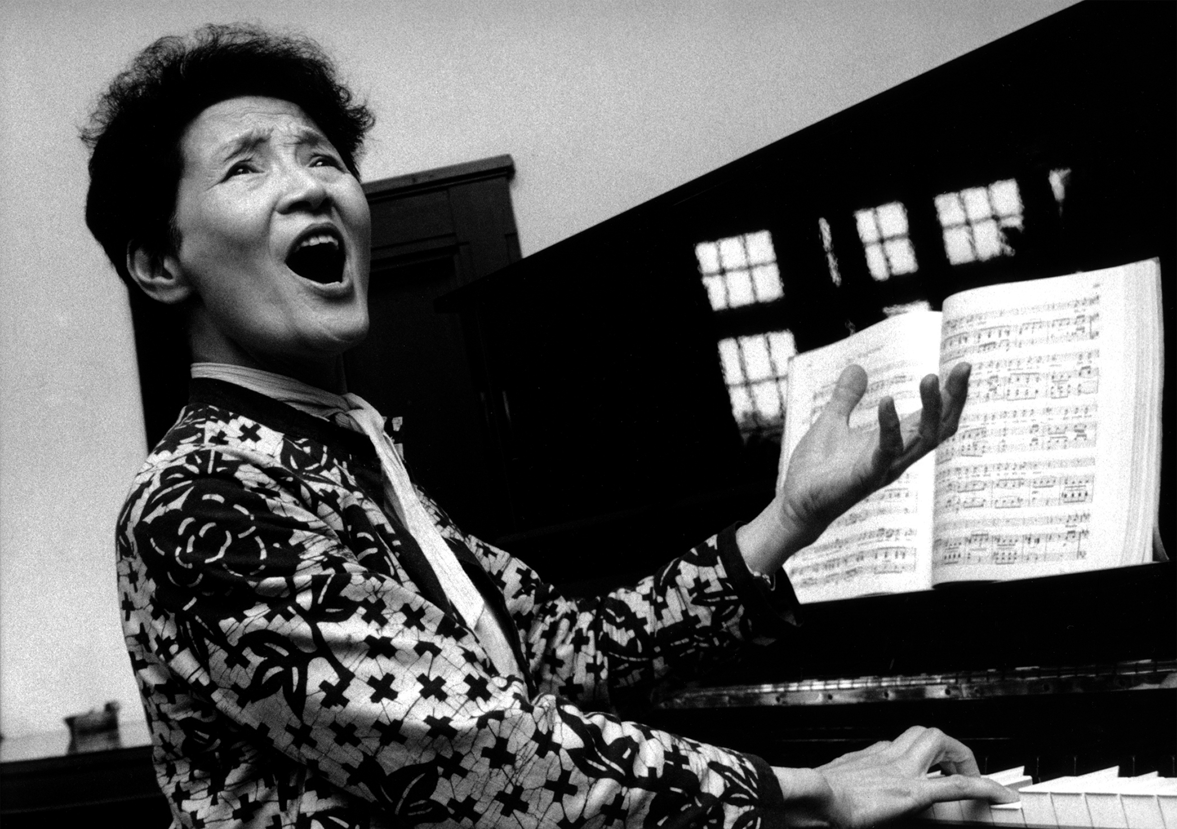 A woman sings with intensity as she plays the piano with one hand and gestures with the other. She is sitting angled to the right, so her full face is visible.