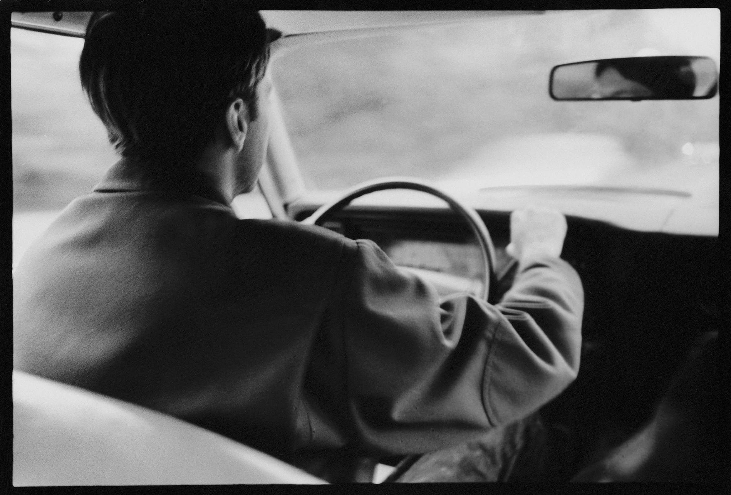 A man wearing a jacket and seen from the back seat is driving an older model car.