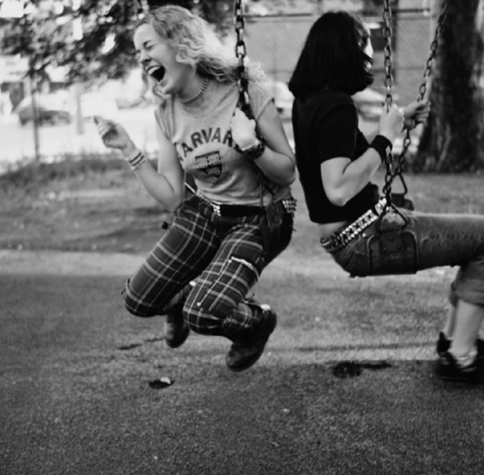 Two teen girls are on swings. One girl has long blond hair and is laughing really hard with her eyes closed and mouth wide open. The other girl has brunette hair and is seen from the side facing away from the camera.