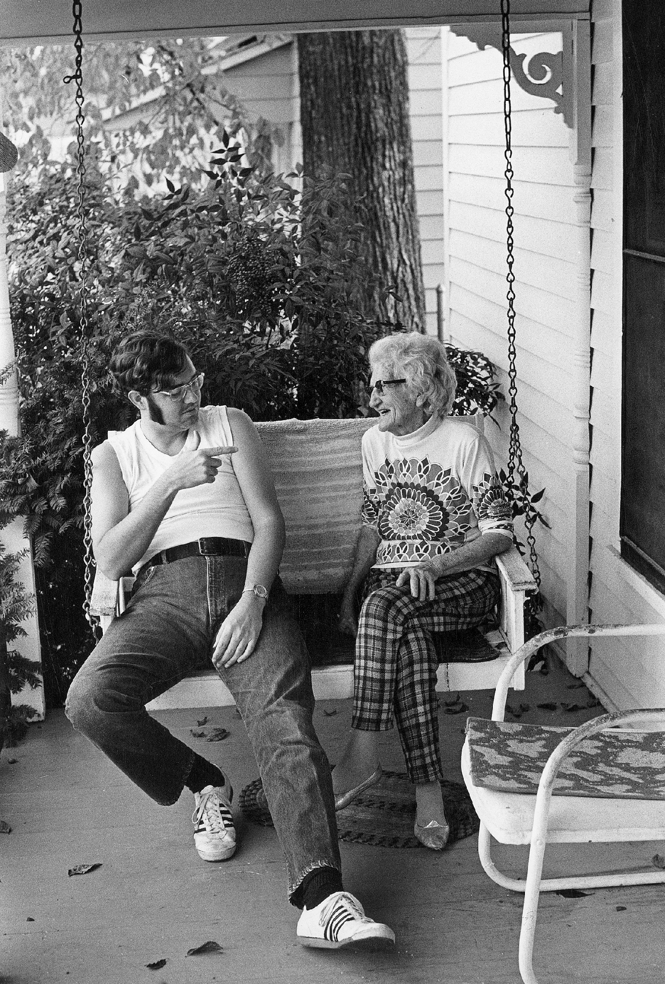 A young man with long, thick sideburns and glasses wearing a white sleeveless T-shirt, jeans, and “track shoes” circa 1970 and an old woman in a sweater and plaid pants sit and talk on a suspended swing on a porch. He points at her while she smiles.