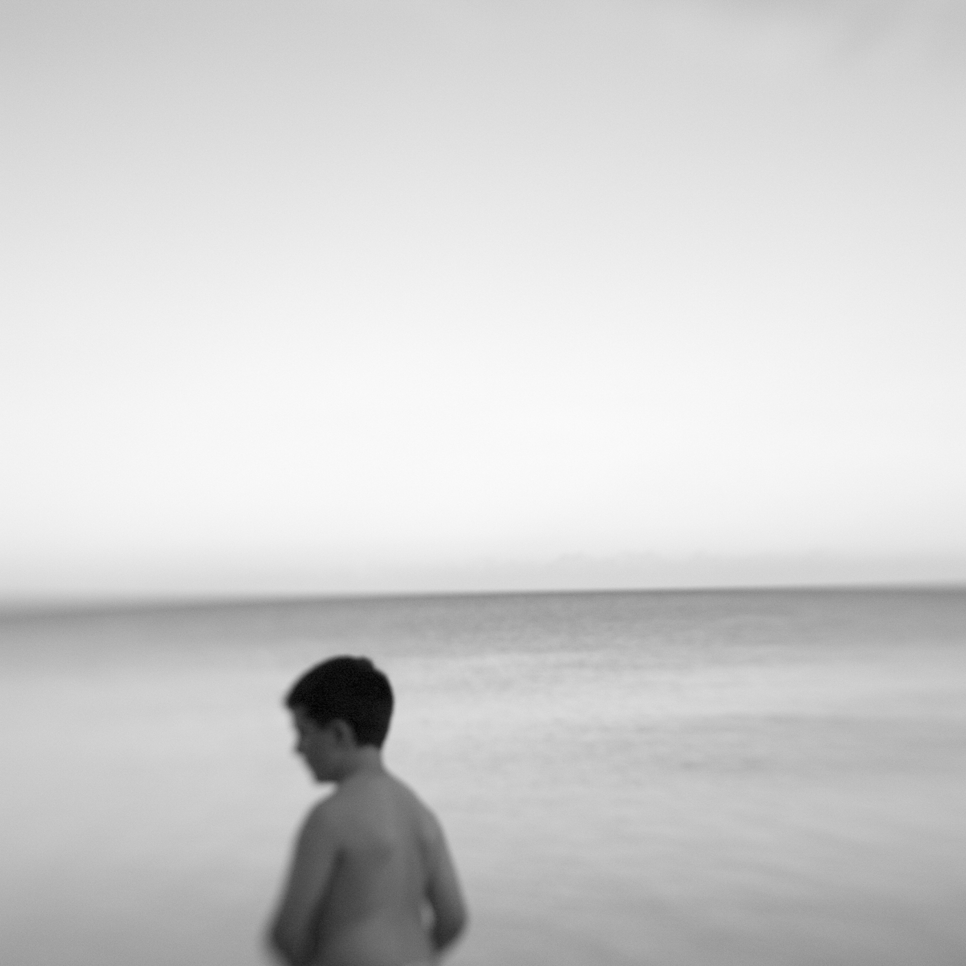 The top half of a shirtless boy in the foreground with a body of water to the horizon in the background.