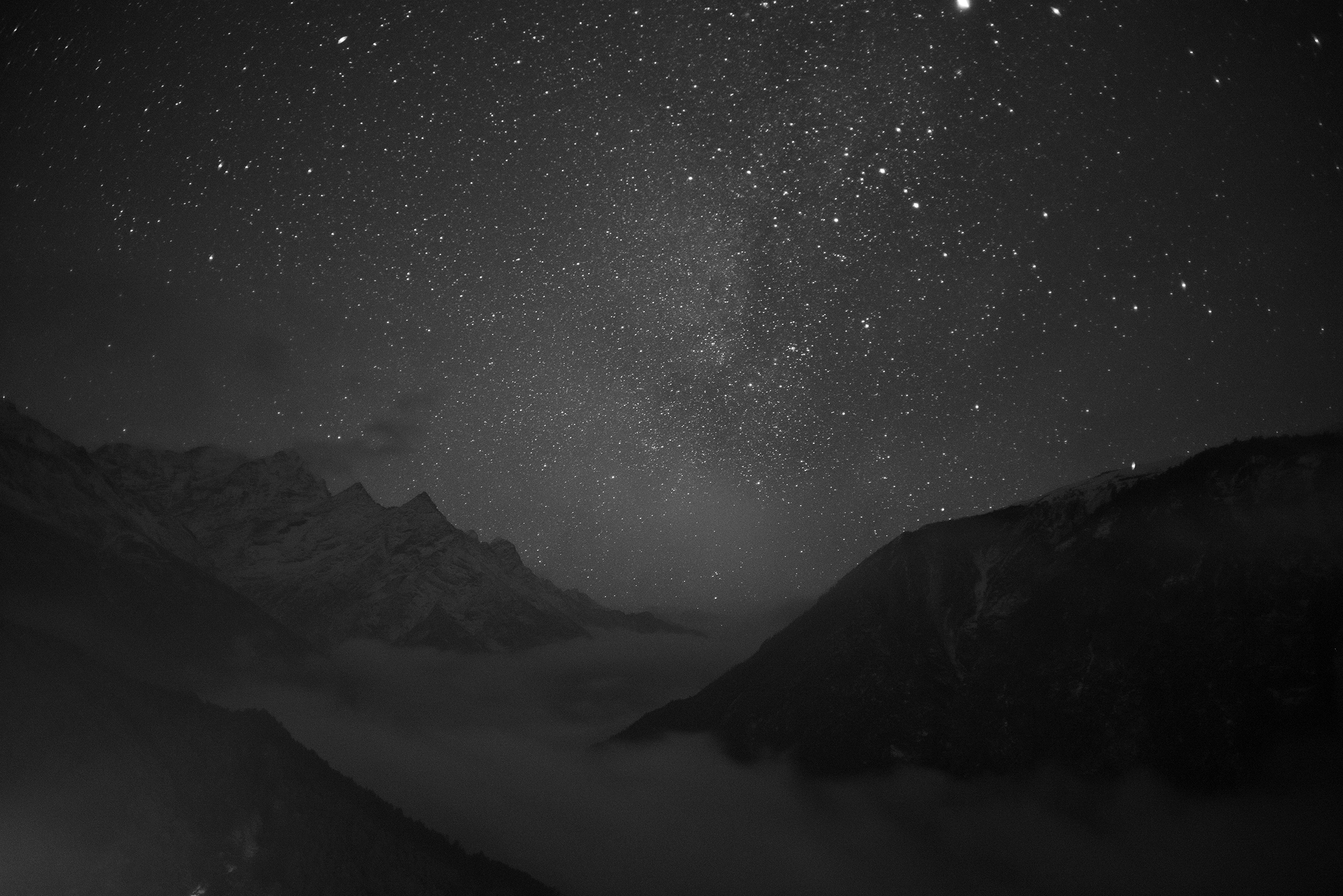 The tops of several mountains with  clouds between them and below the peaks on a very starry night.