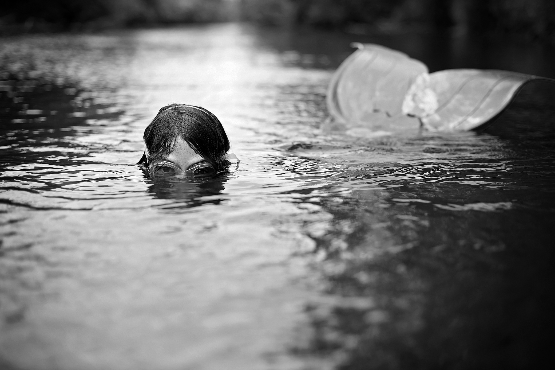 A young girl wearing swimming goggles is swimming in a river. Only the top part of her face and the fluke of a mermaid’s tail are visible above the sunlit water.