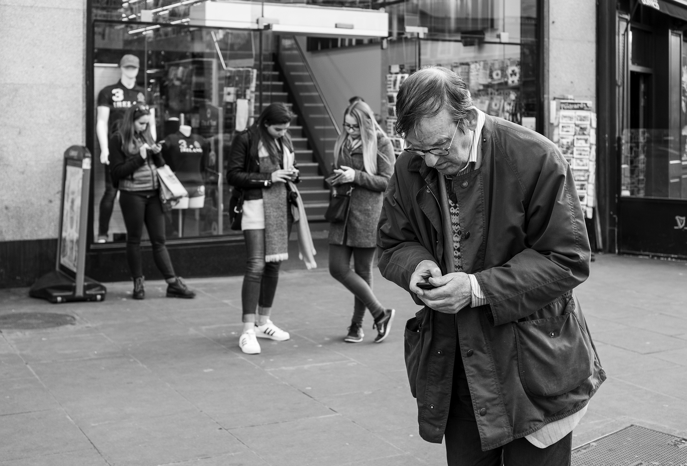 Four people stand near a storefront on a city sidewalk and each one is looking down and interacting with their phone.