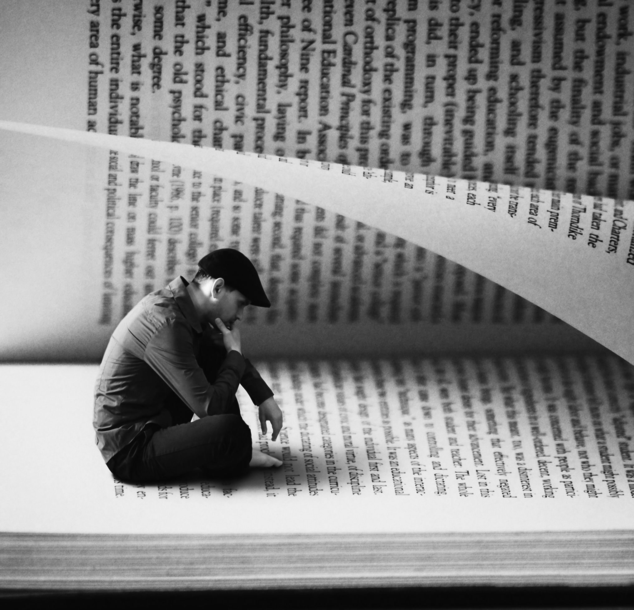 A surreal image of a man in profile sitting inside of an open book with a page curling over on top of him. He is sitting on a book page and looking down as though reading. He is wearing a long-sleeved, button-down shirt, pants, and a flat cap.