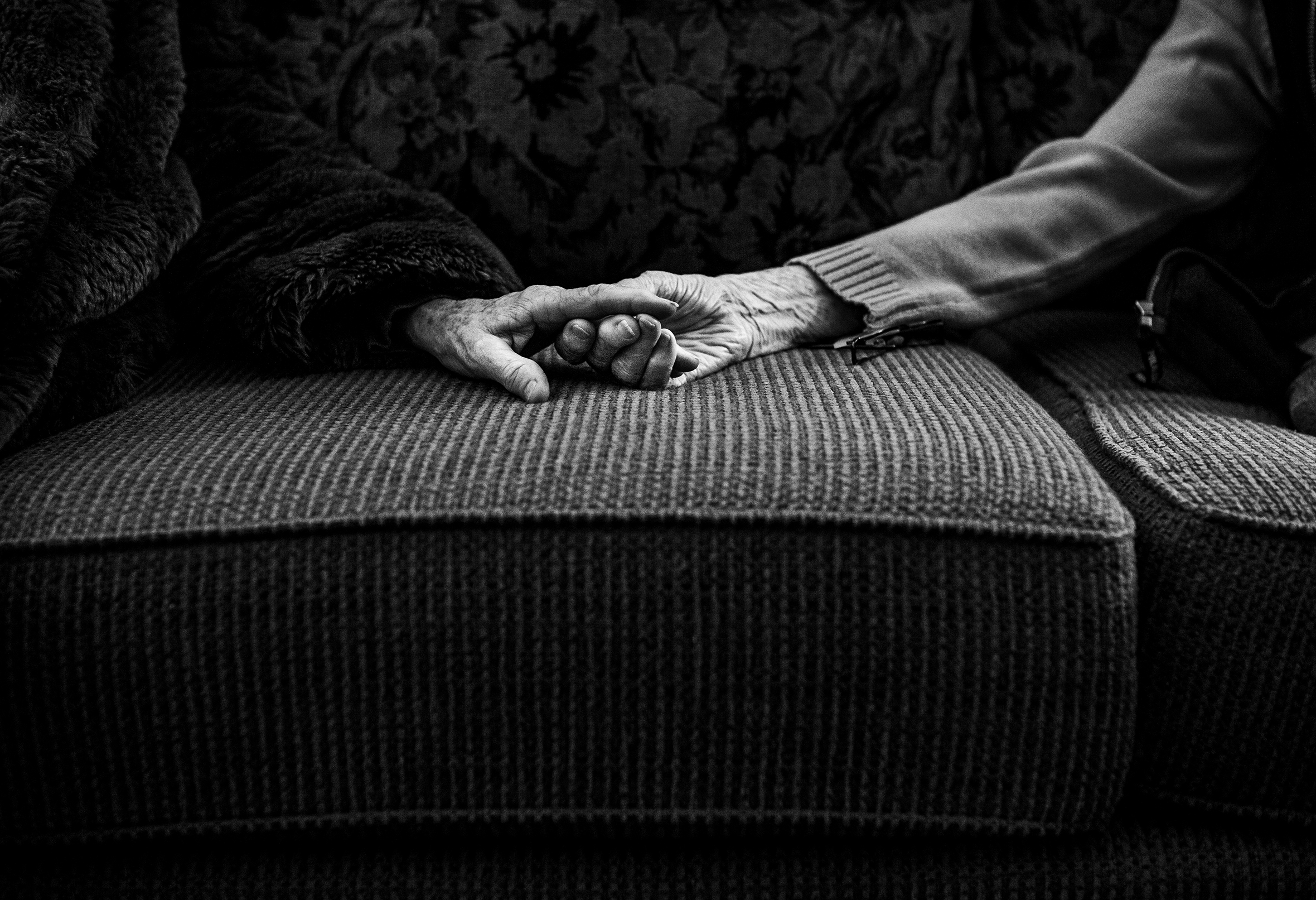 Two elderly people holding hands on a sofa with a close-up on the hands.