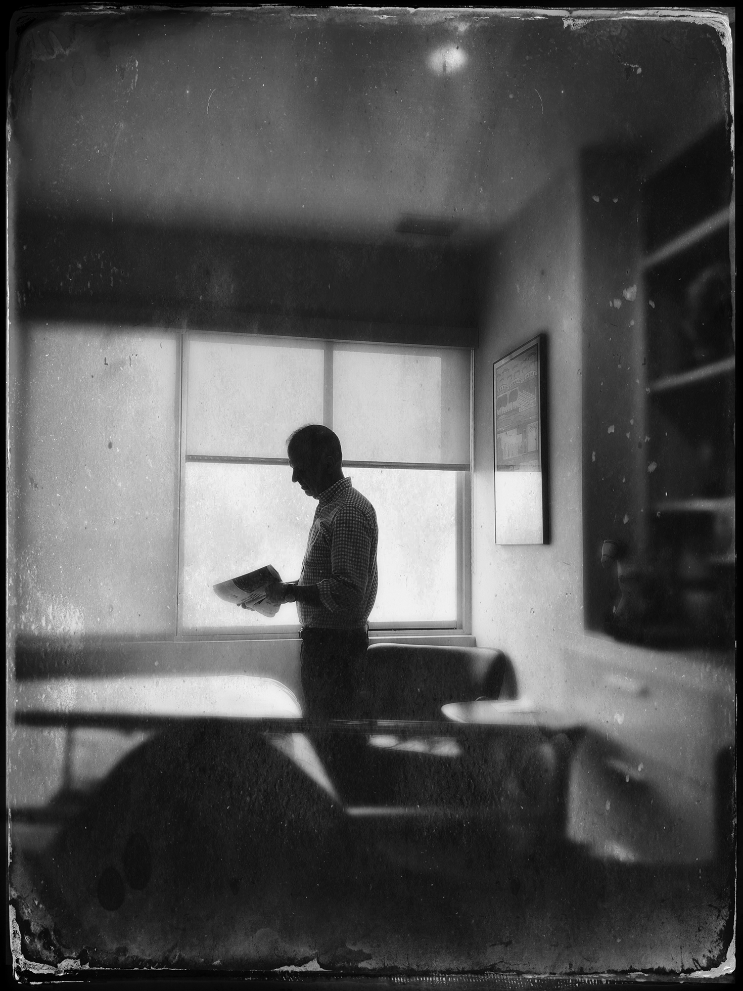 A male teacher stands silhouetted against the window of an empty classroom. He is looking down at papers in his hands.