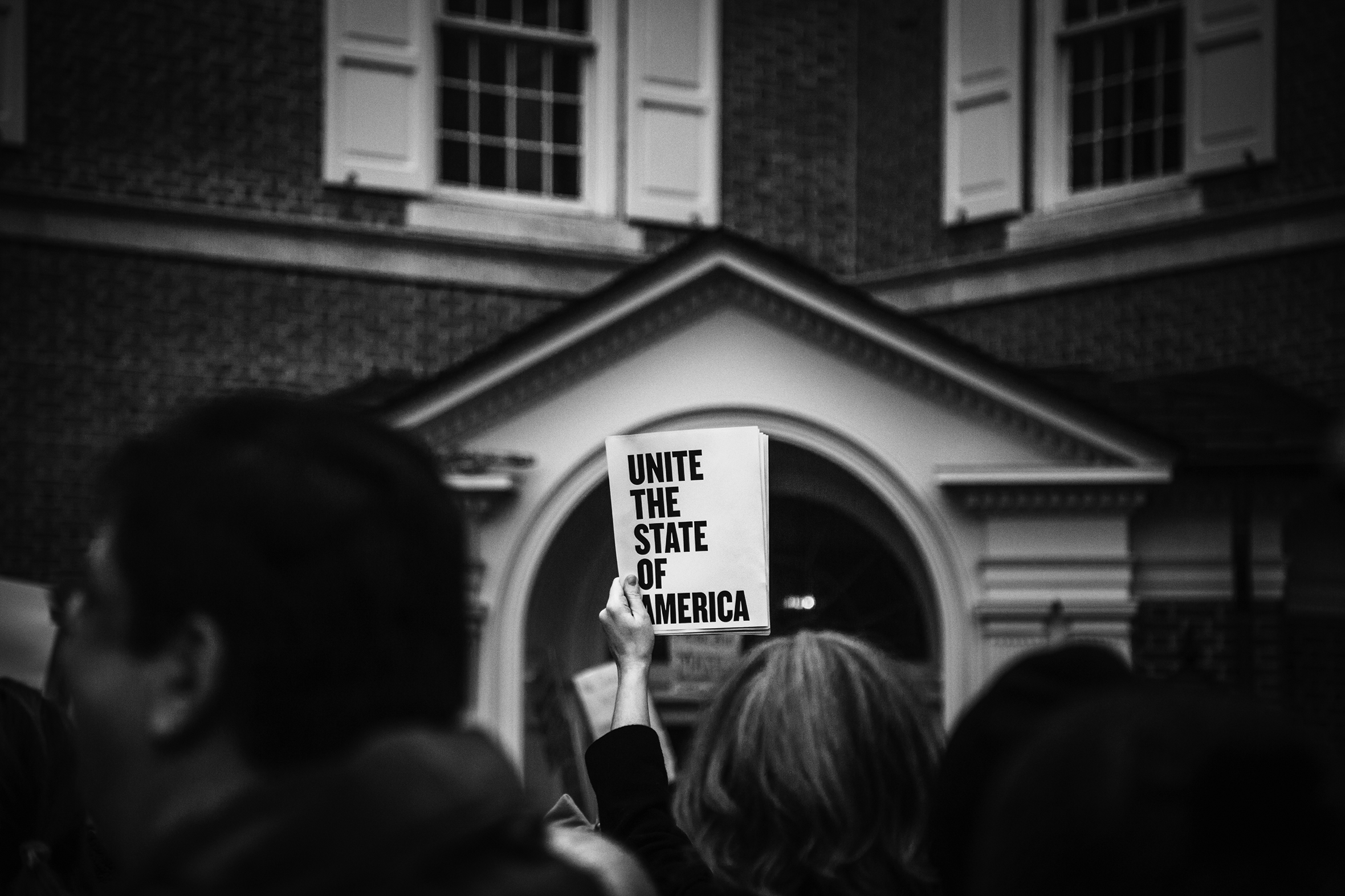 A crowd of protestors is in front of a building. The camera is focused on one sign that stands out and says Unite The State Of America.