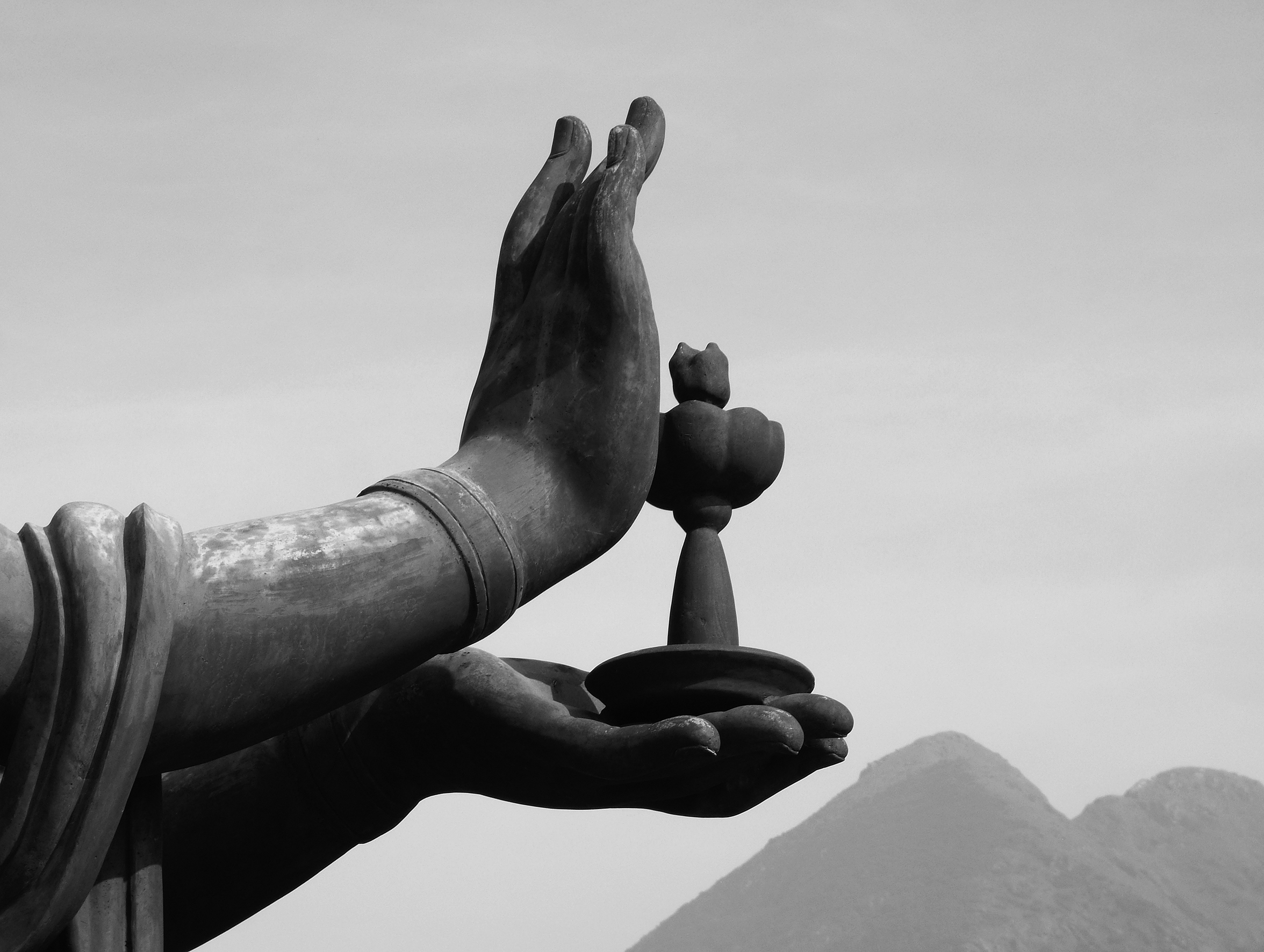 A close-up of a statue’s hands holding what appears to be a lotus. Mountaintops are just peeking from the bottom-right corner of the image.