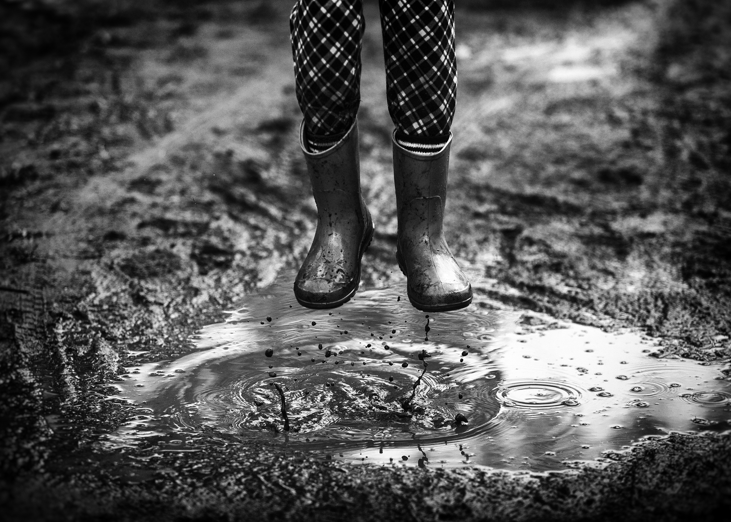 A close-up of a child’s legs with the pants tucked into rain boots taken mid-jump over a mud puddle.
