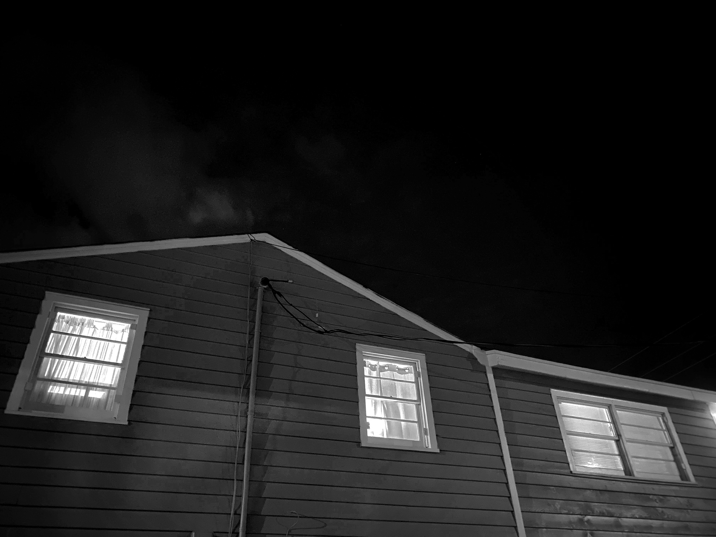 An upward shot of the side of a one-story house at night. The lights are on and the curtains are drawn.
