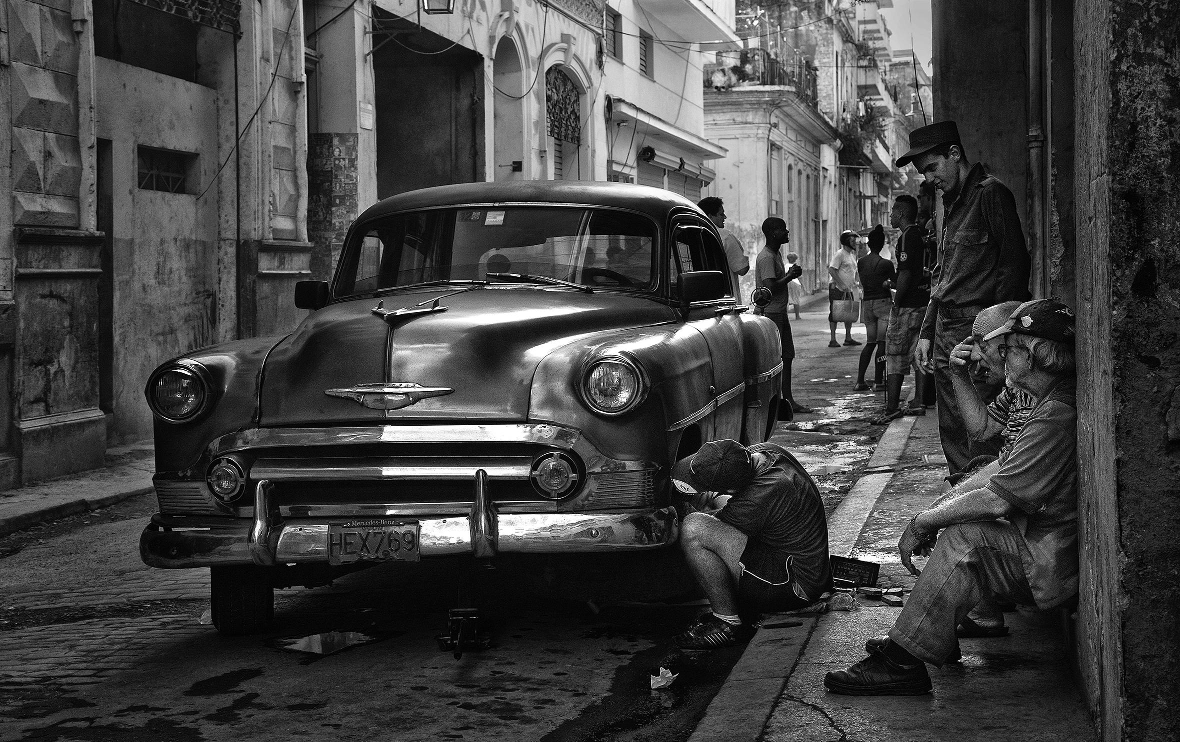 A young man is working on a vintage car with a Cuban license plate parked on the street. A couple of old men are watching from the sidewalk as people go about their daily lives in the background.