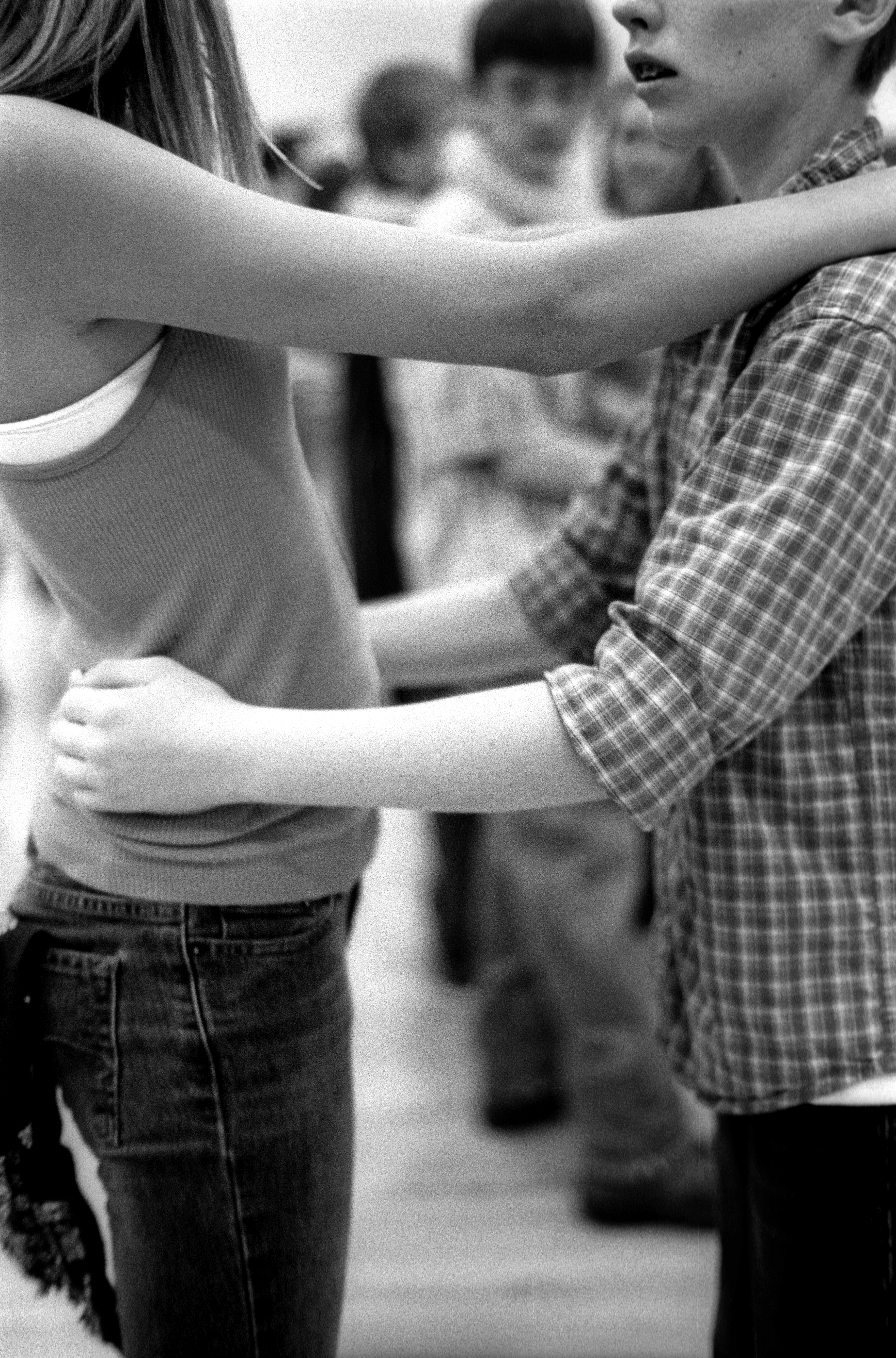 A teenage boy and girl are slow dancing. He has his hands on her waist and her forearms are resting on his shoulders, and there is distance between them. In the background, blurred, are other teen couples dancing.
