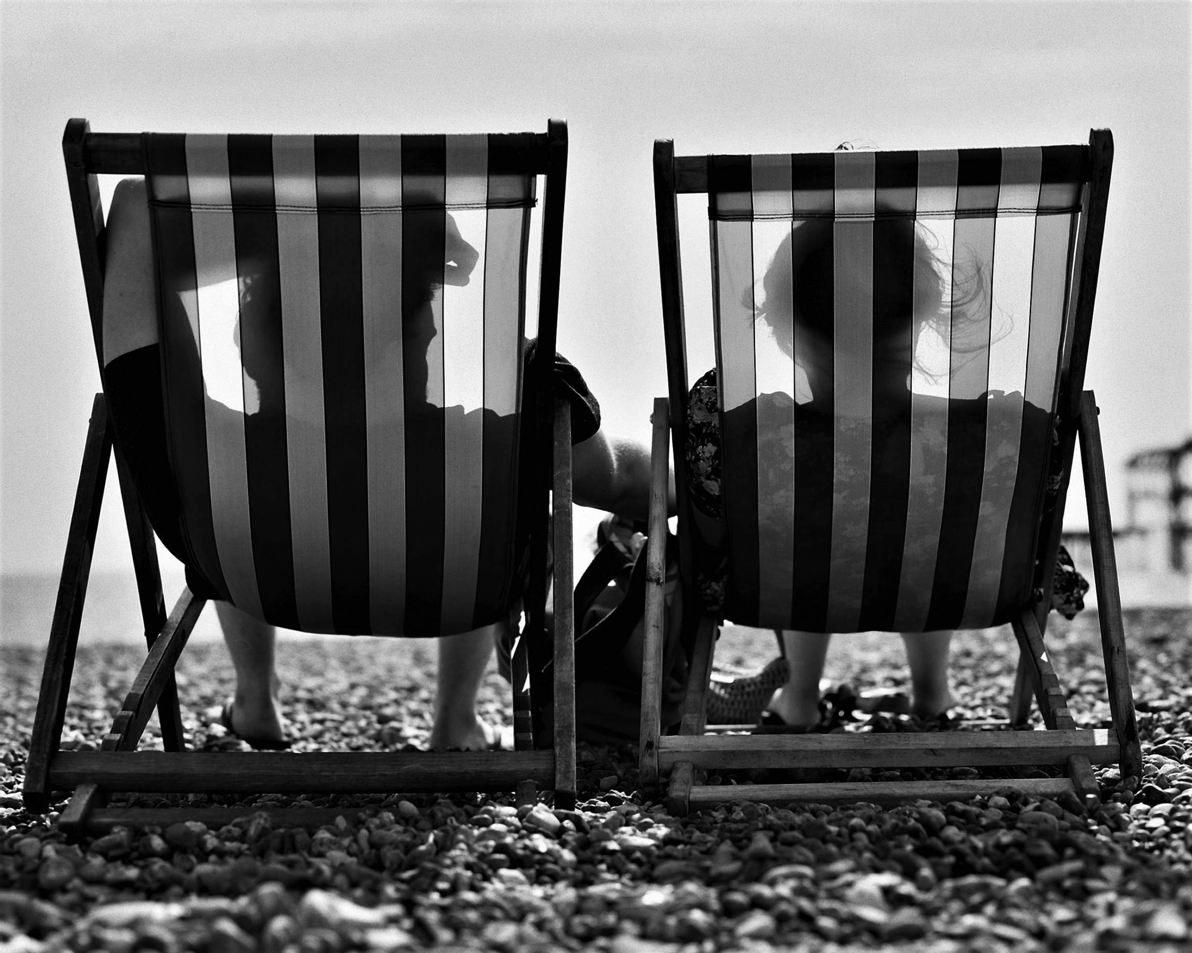 A couple sitting on a rocky beach in matching striped beach chairs, facing the water.