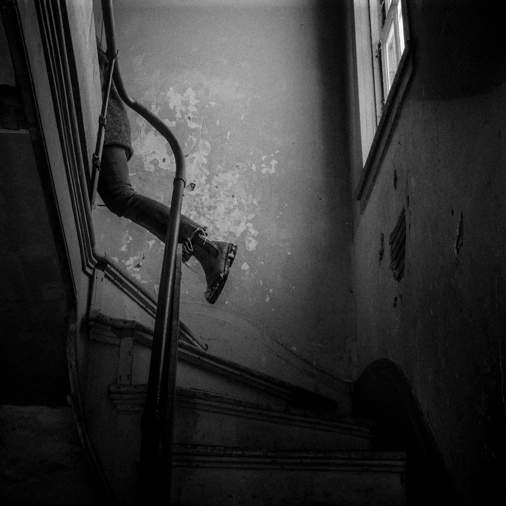 A glimpse of a person’s left leg as they climb stairs and round the next flight to the left. They are wearing a wool coat and frayed jeans and a boot that has a bit of snow on the side. The stairwell looks well used and in need of paint.