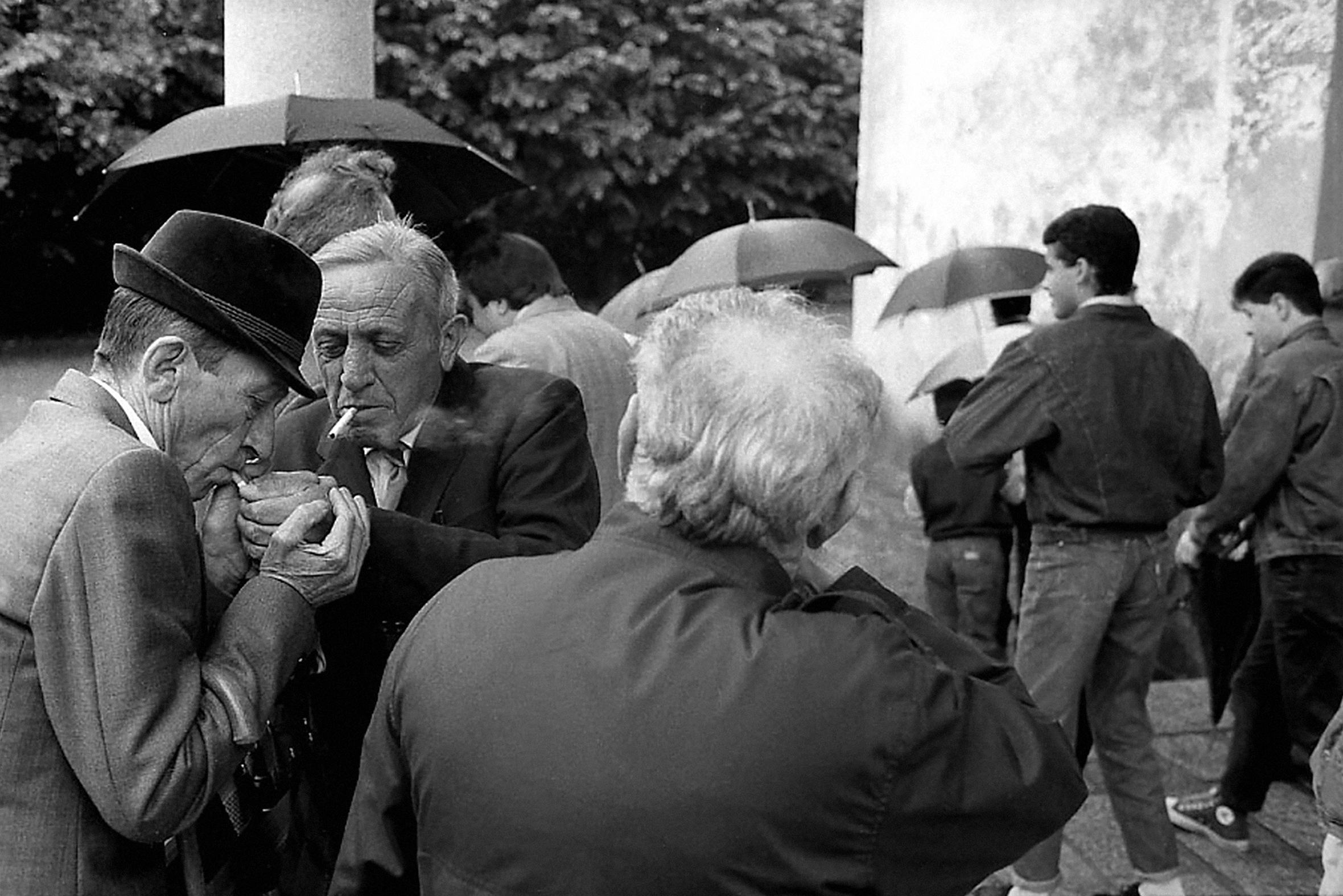 The backs of people walking out of a building wearing suitcoats and light jackets, some with umbrellas. On the left are two older men: one is lighting the other's cigarette.