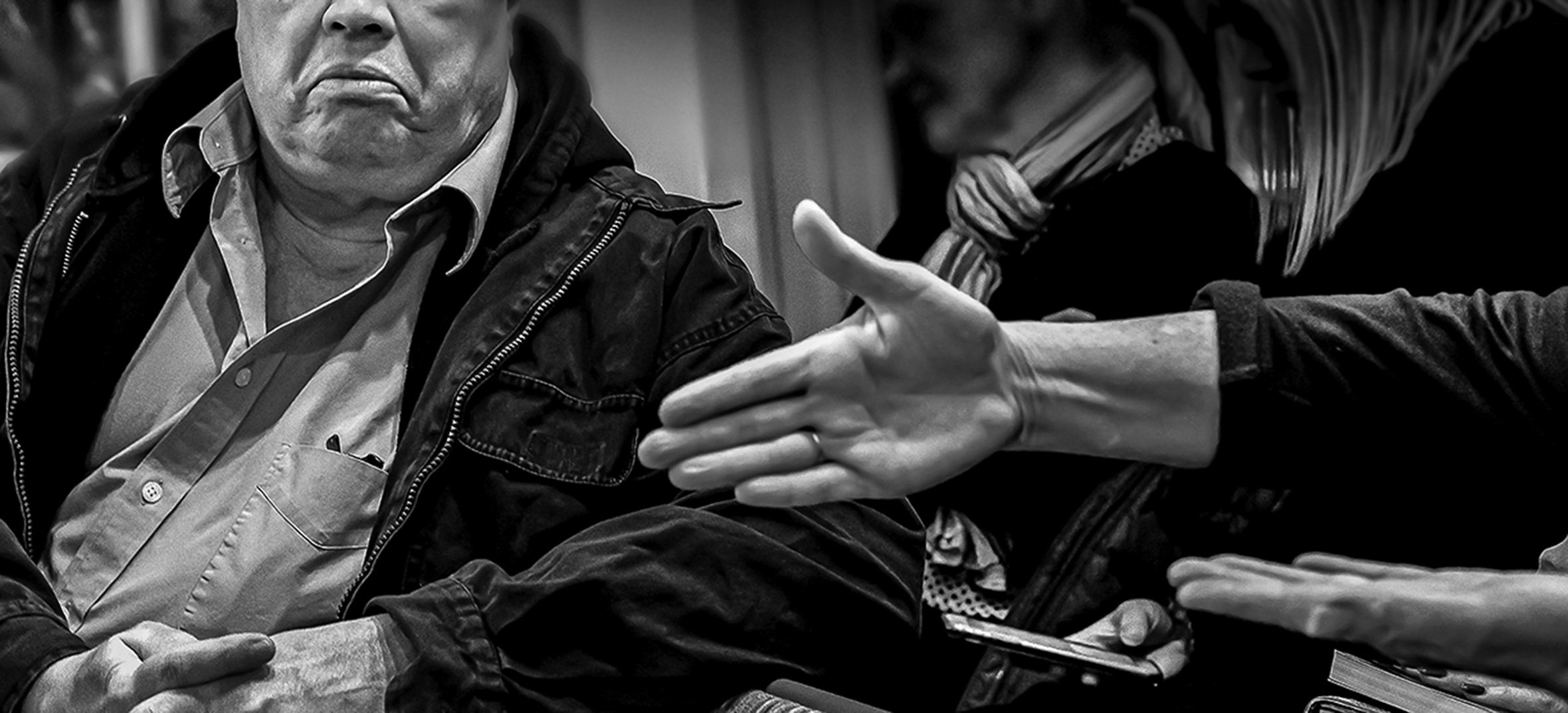 A man on the right has extended his hand across a table for a handshake with a man on the left. The man on the left is sitting with his elbow on the table and the bottom part of his face is visible. His hands are clasped and his mouth is downturned.