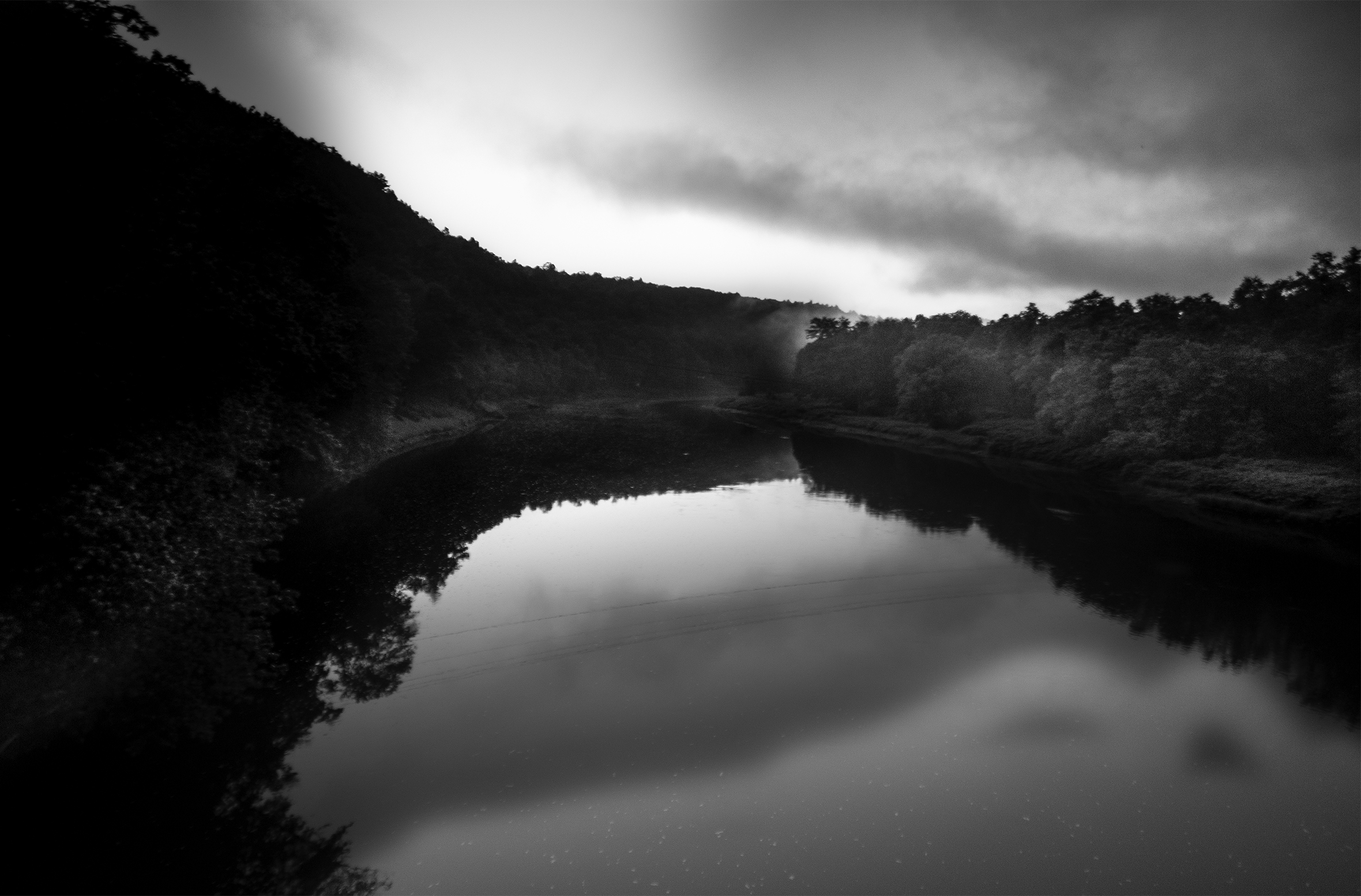 A wide, placid river curving off into the distance. The left bank is a continuous ridge with vegetation and the right bank is flat with trees along it.