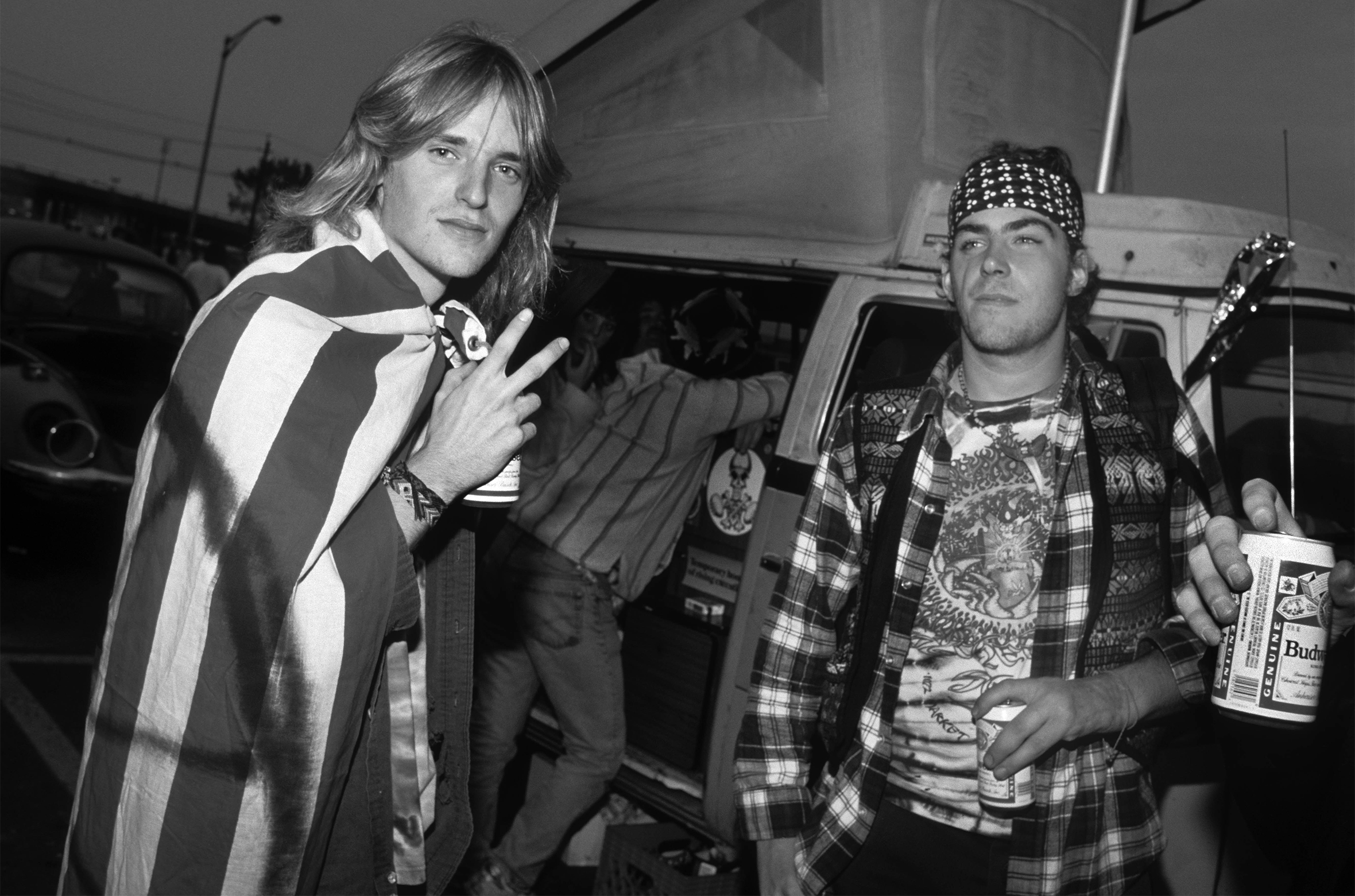 Young men stand in a parking lot in front of a van drinking Budweiser beer. The man on the left with long blonde hair and a flag draped over his shoulders looks into the camera and flashes a peace sign. The man on the right has a bandanna on his head.