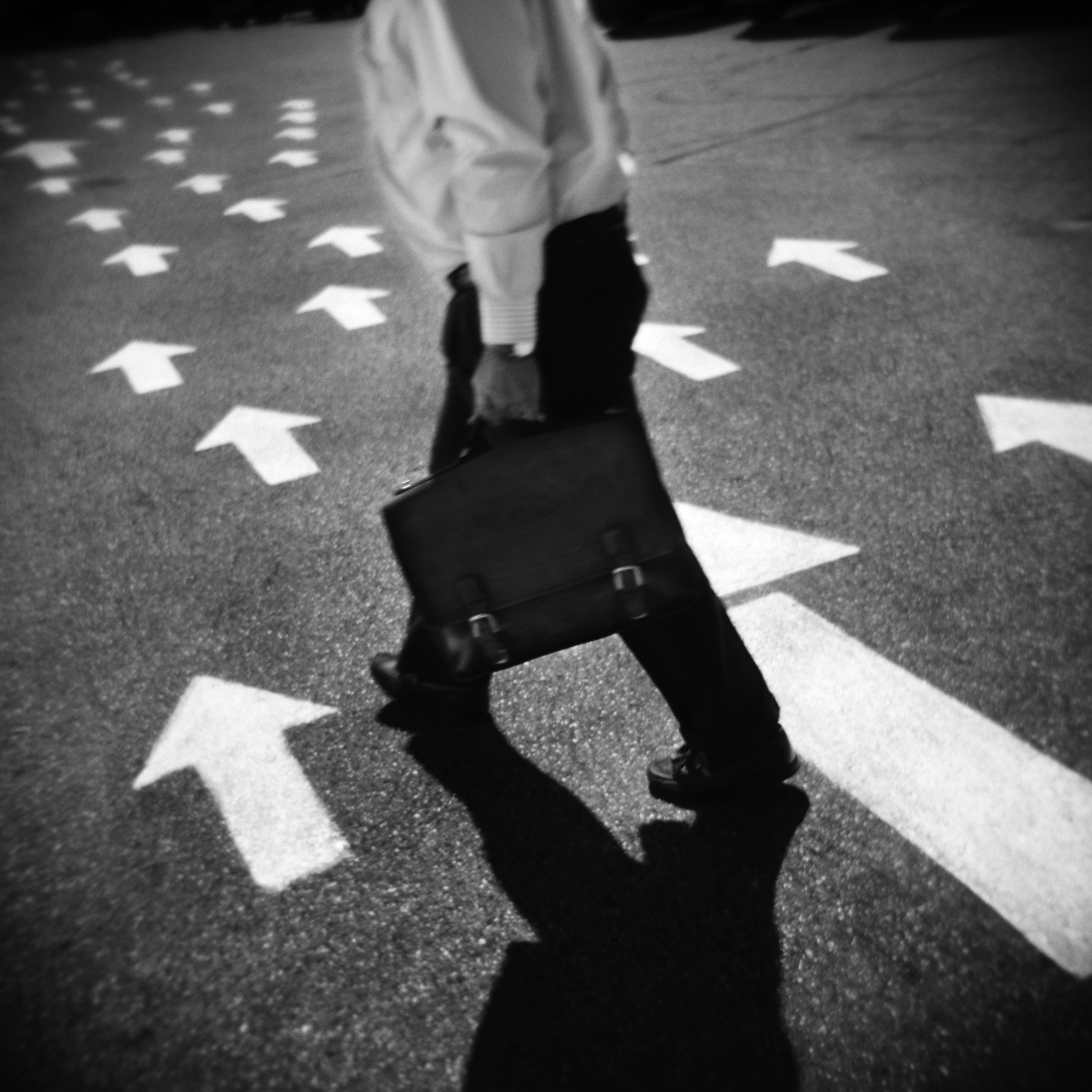 A man shown from the neck down in business attire carrying a briefcase is walking on pavement that has white arrows pointing in the direction he is walking.