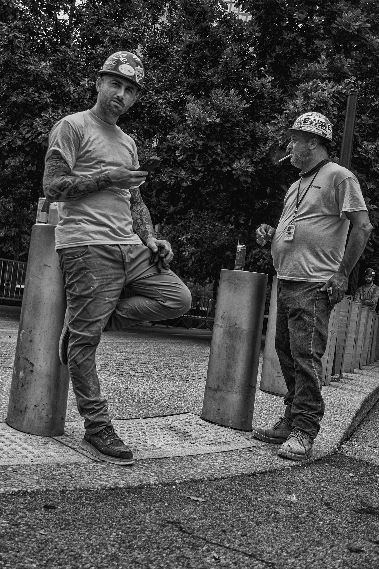 Two men in work clothes and hardhats take a break by metal bollards that they have placed canned beverages on top of. One man with sleeve tattoos leans against a bollard while holding a smartphone. The other man has a cigarette in his mouth.