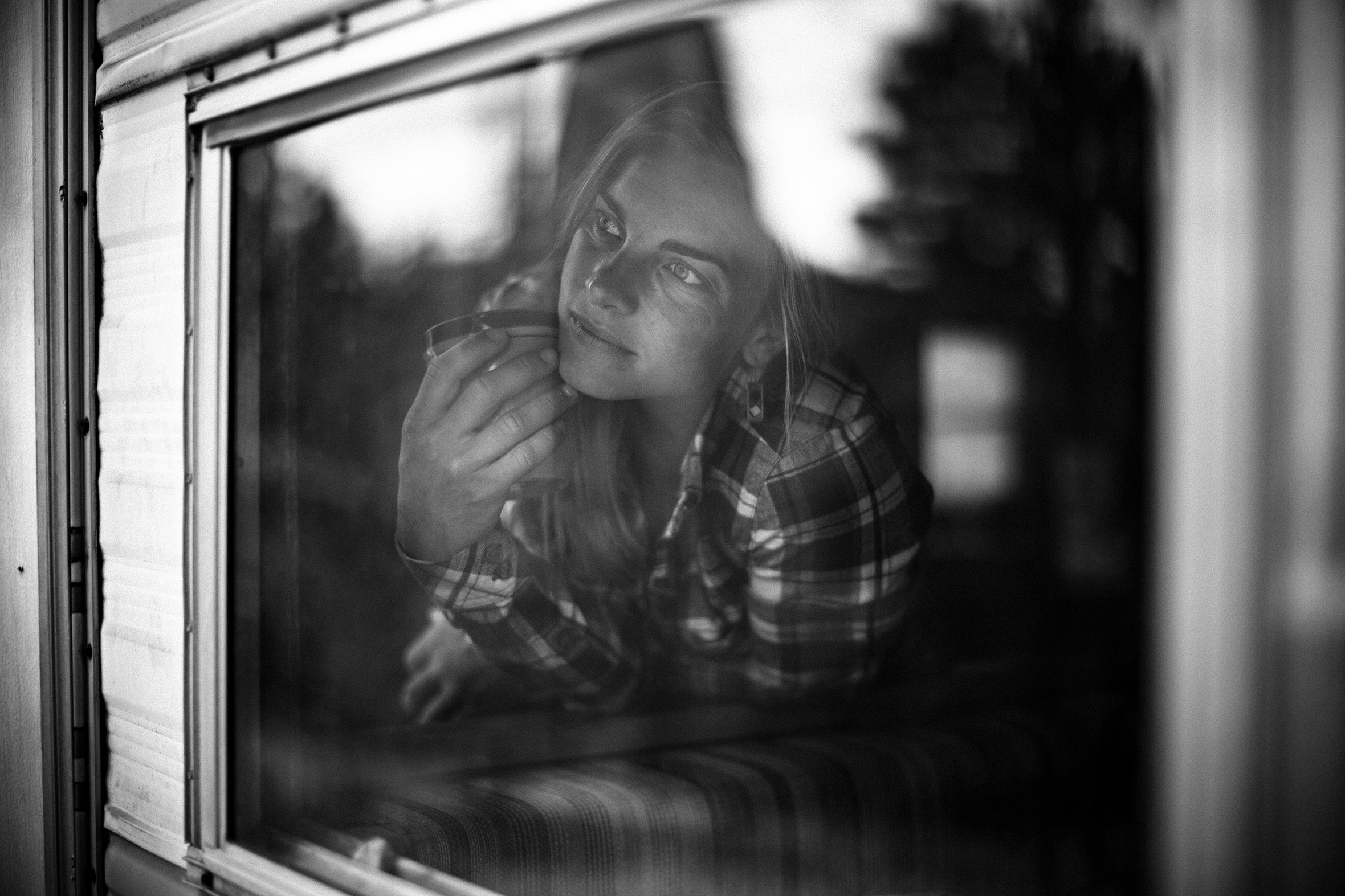 A woman leans forward and looks out of what appears to be the window of an RV. She is wearing a flannel shirt and holding a cup near her face.