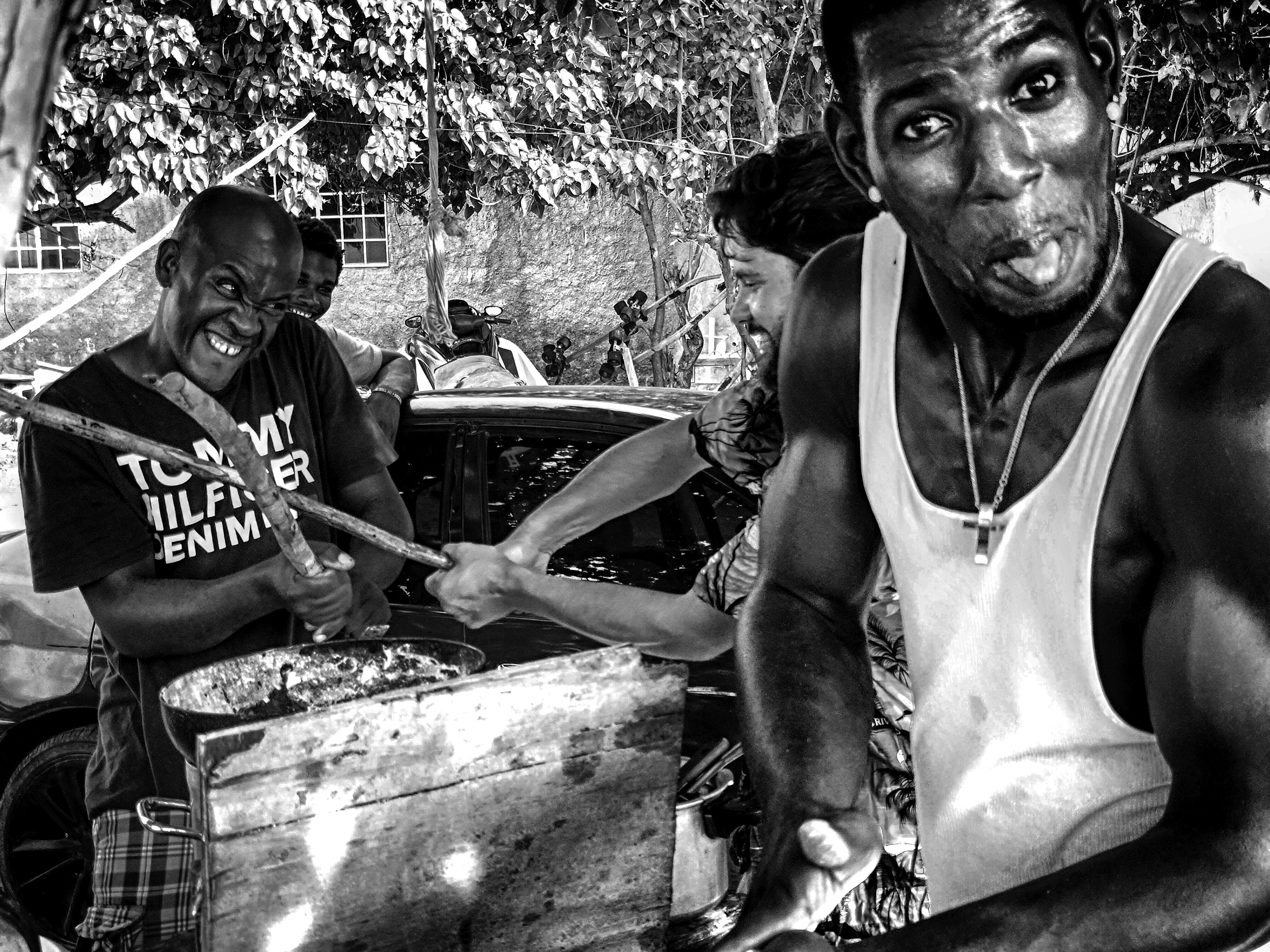 Three men are outside on a street: all are smiling and playful. Two are play fighting with sticks as the third looks into the camera and sticks out his tongue.