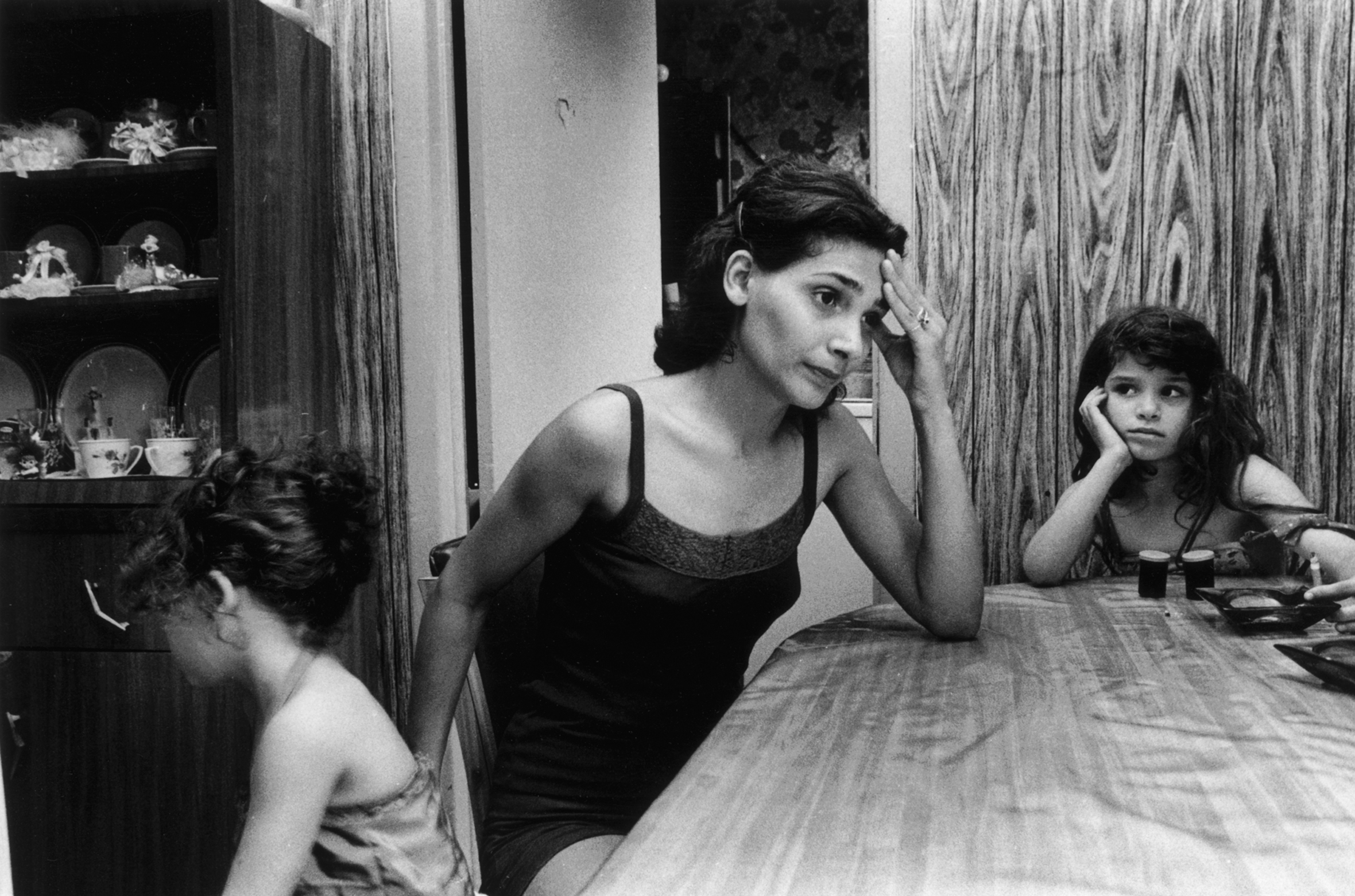 A frustrated-looking woman sits at a dining-room table with two young girls. The woman has her left elbow on the table with her hand on her forehead. One girl faces away, while the other rests her head in her hand and looks at the woman.