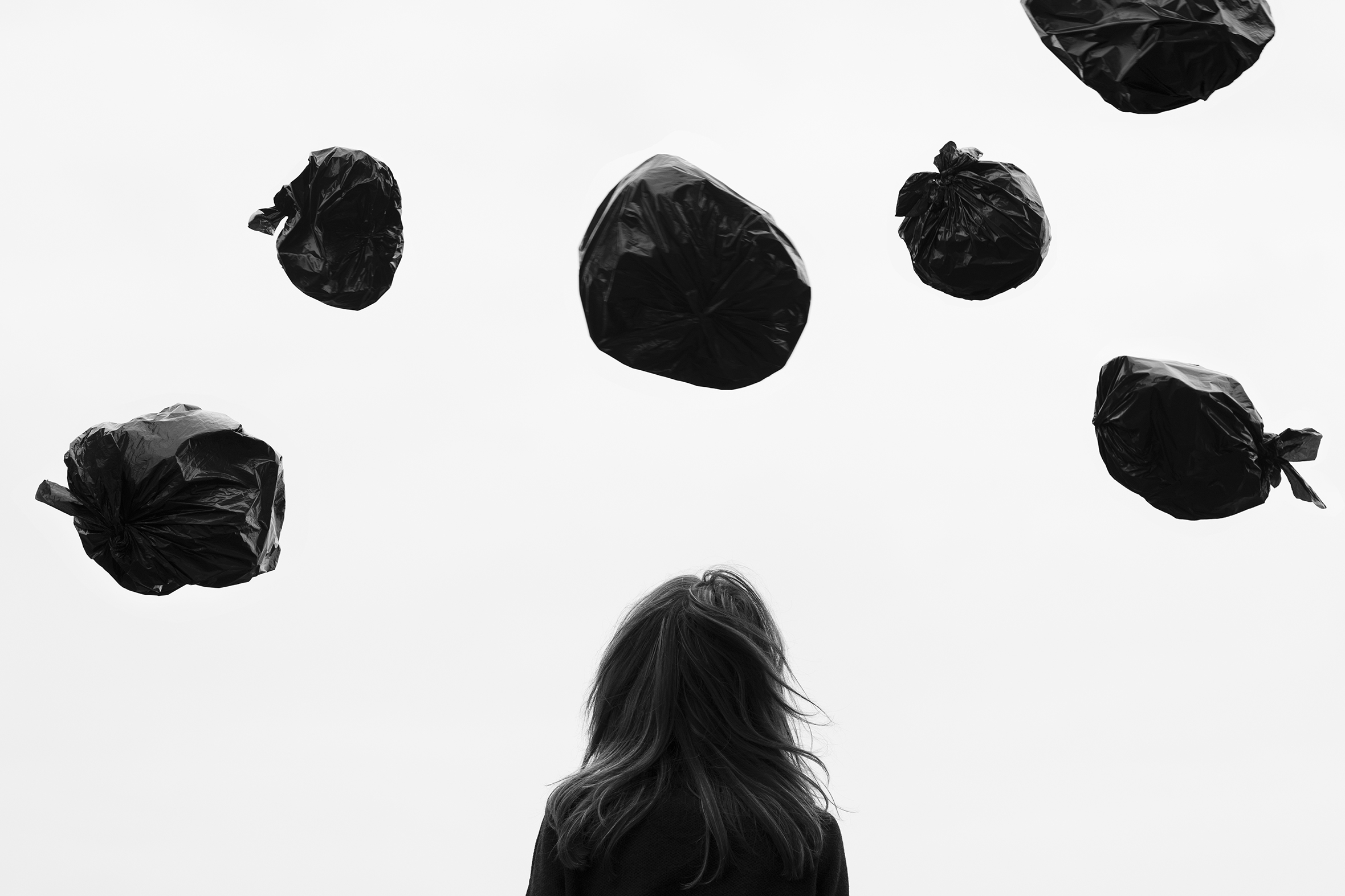 The top half of a woman with long hair seen from the back looks up at six trash bags thrown up in the air.