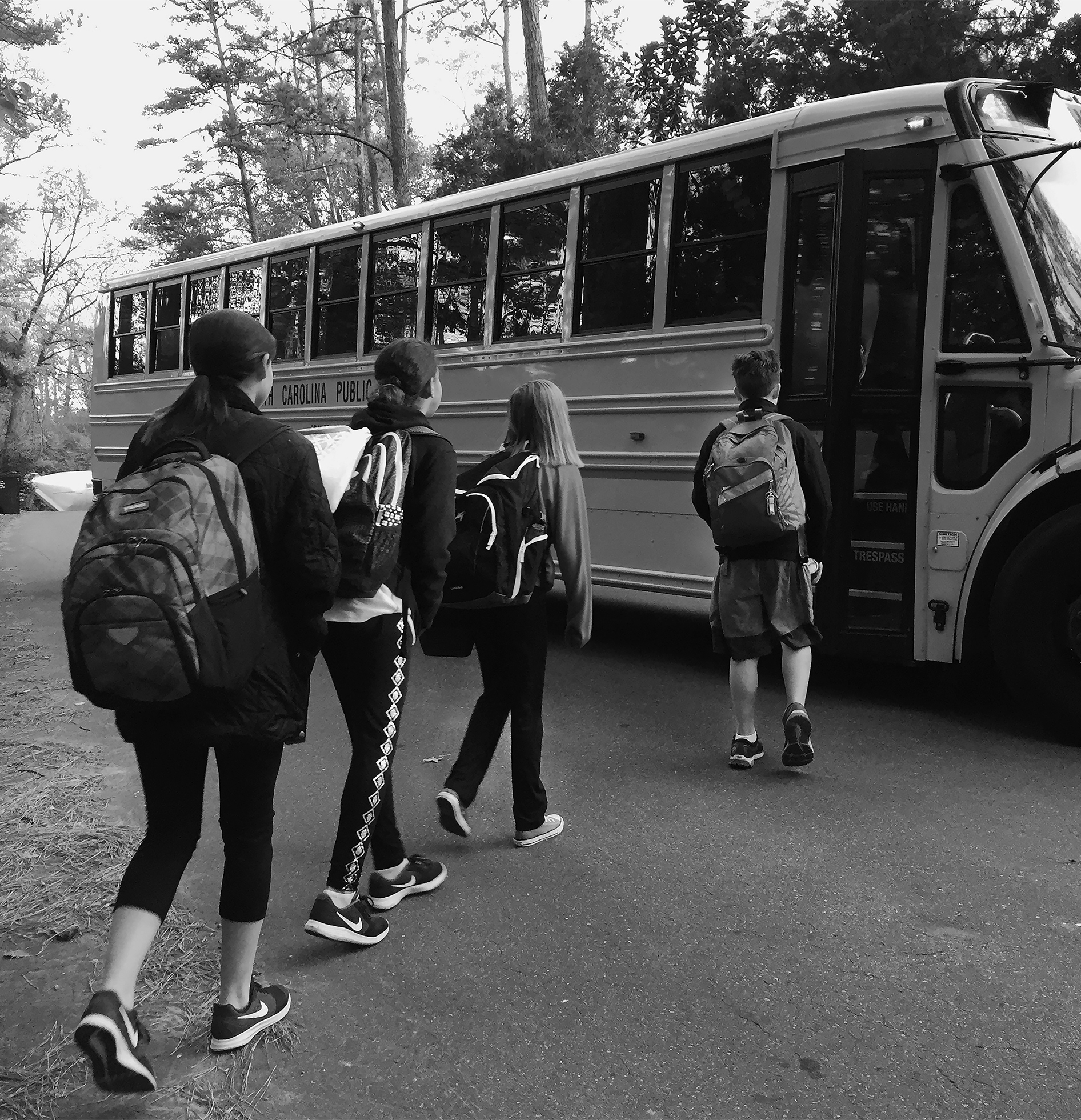 Four children in jackets with backpacks on a rural road walk toward a school bus to board it.