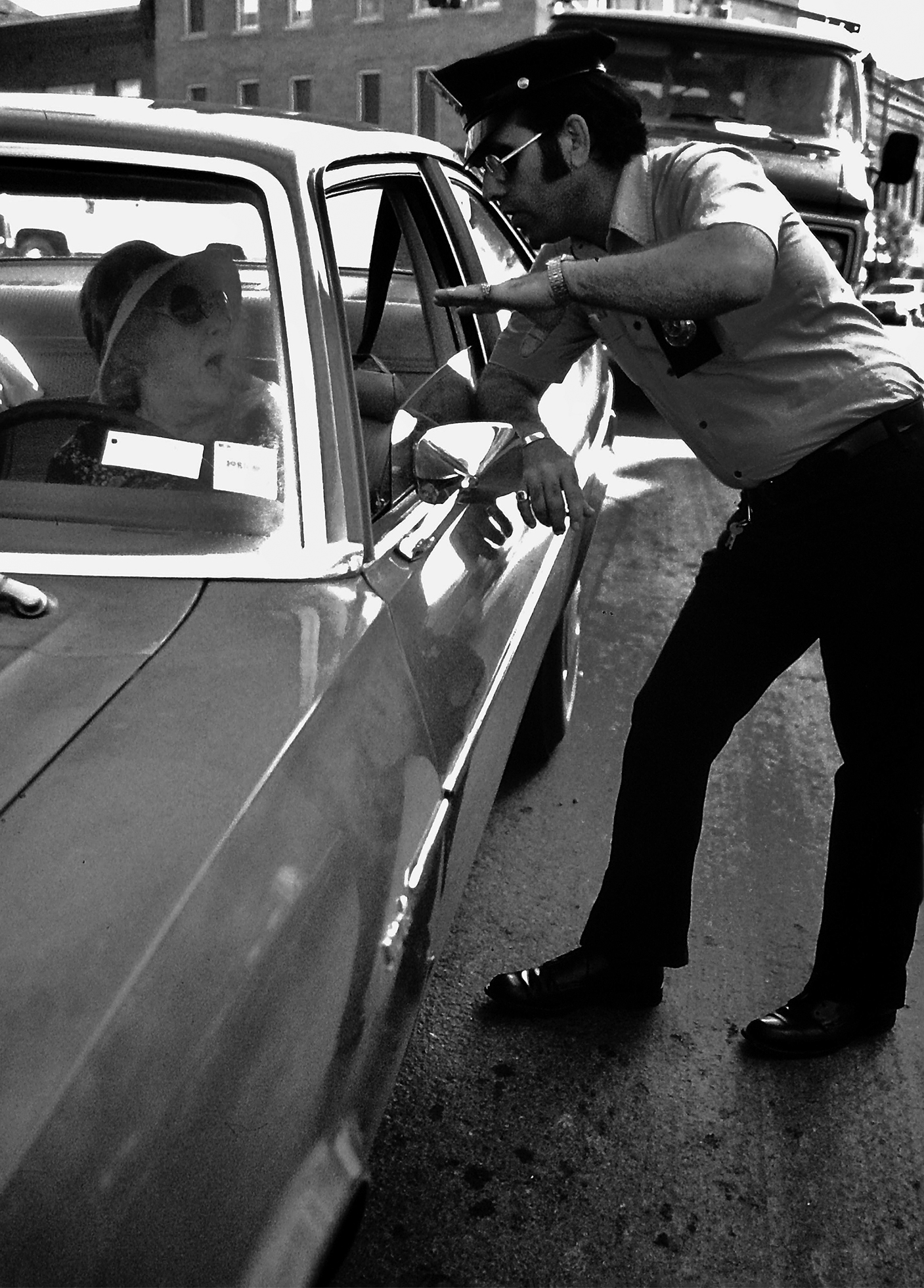 A police officer with sideburns and dressed from the 1970s speaks with an older woman in her car through the driver’s side window as he leans against the car door.