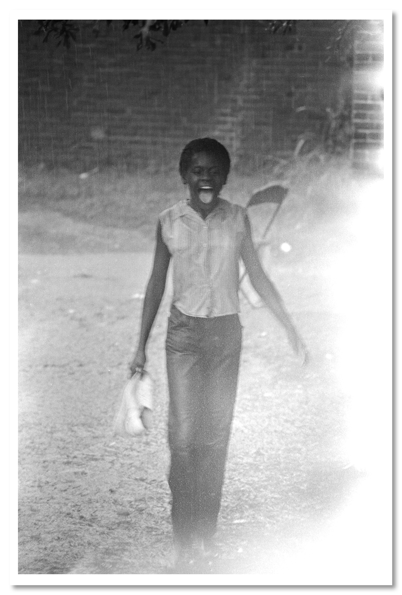 A young woman with her mouth open as rain falls walks toward the camera, her shoes pinched in her right hand. The photo is archived as “Young woman standing in the rain during a civil rights demonstration in Greensboro, Alabama,” and dated July 1965.