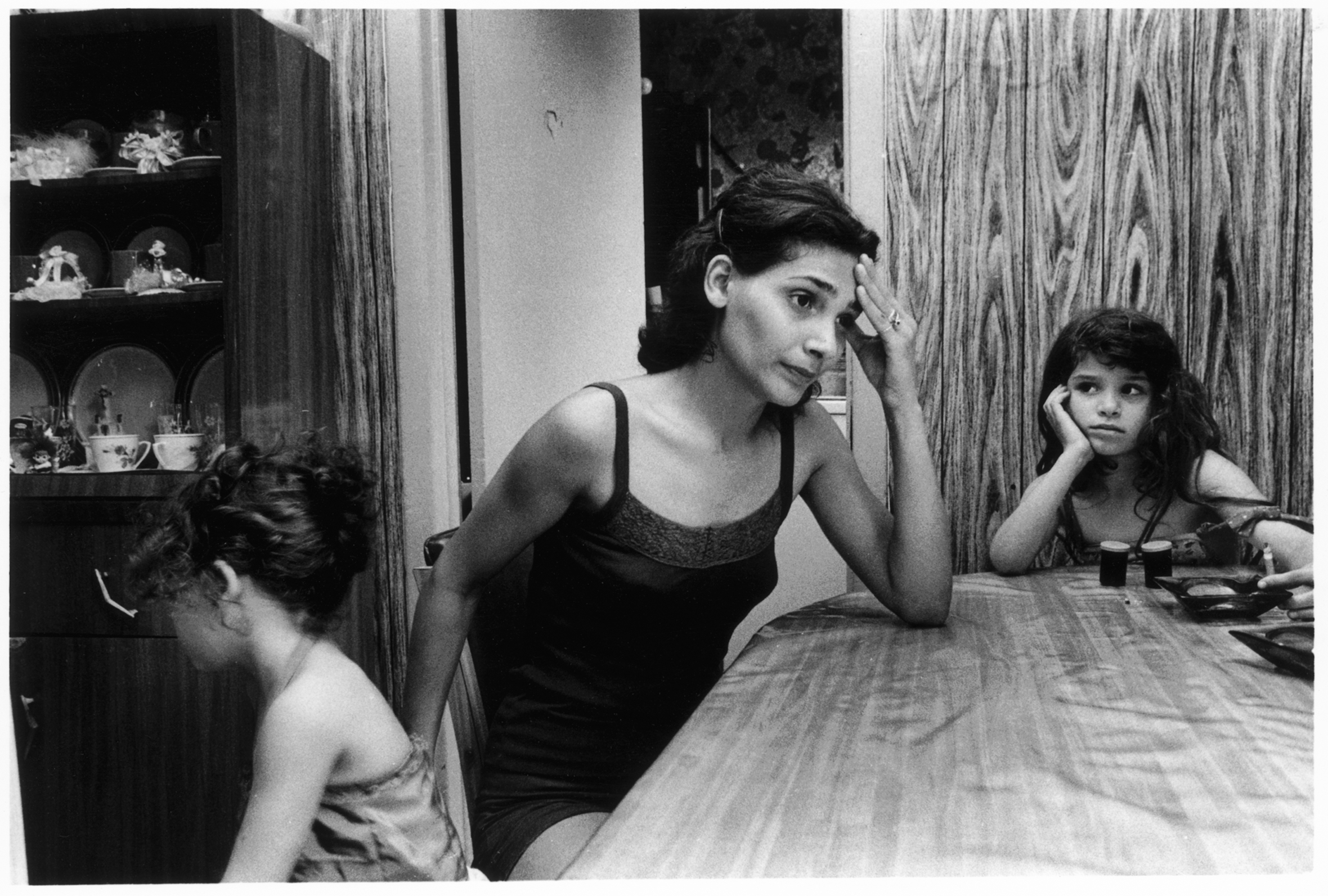 A frustrated-looking woman sits at a dining-room table with two young girls. The woman has her left elbow on the table with her hand on her forehead. One girl faces away, while the other rests her head in her hand and looks at the woman.