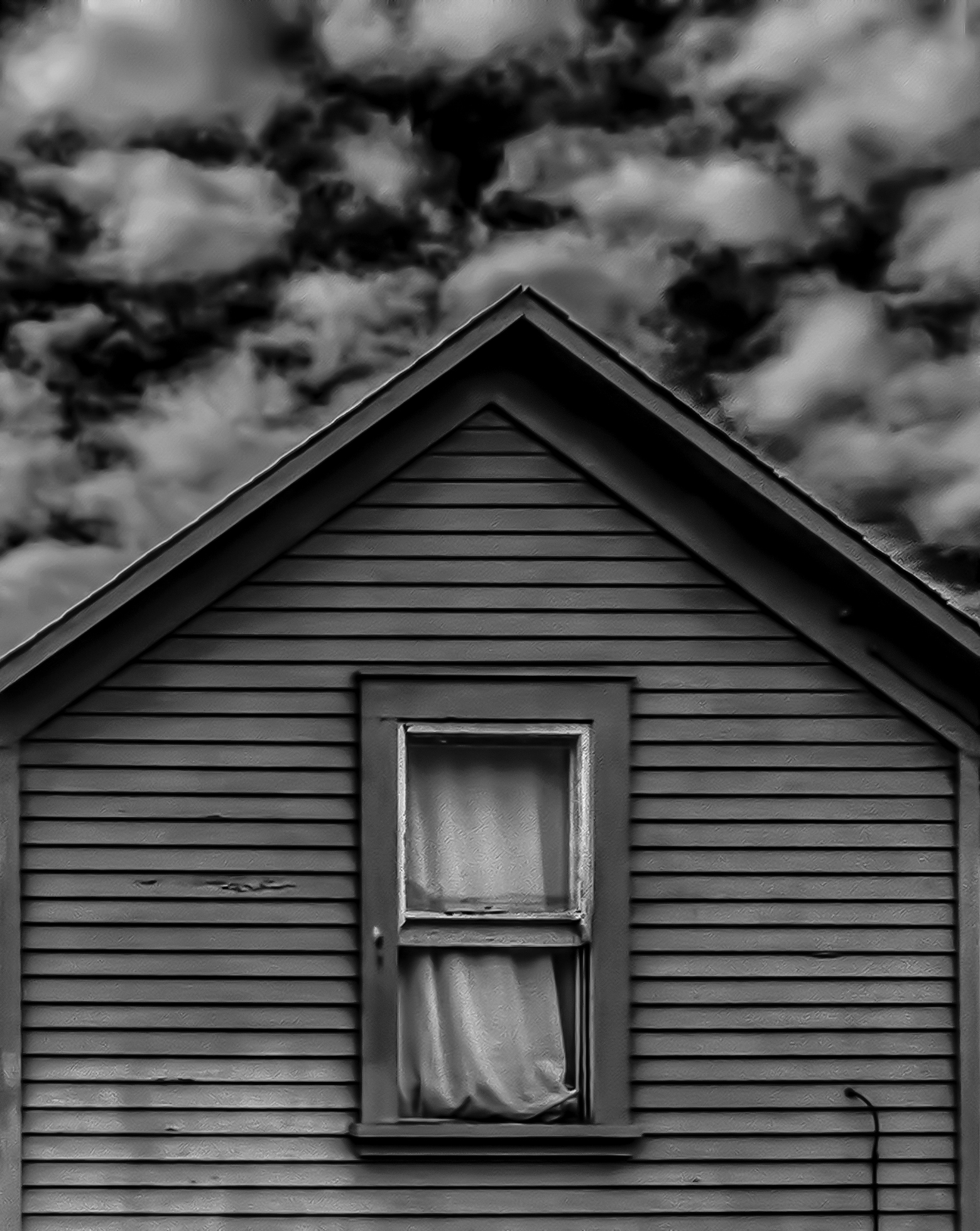 The side of an older, one-story house with horizontal panels, a gable roof, and one single-hung open window with the curtains billowing outside of it.