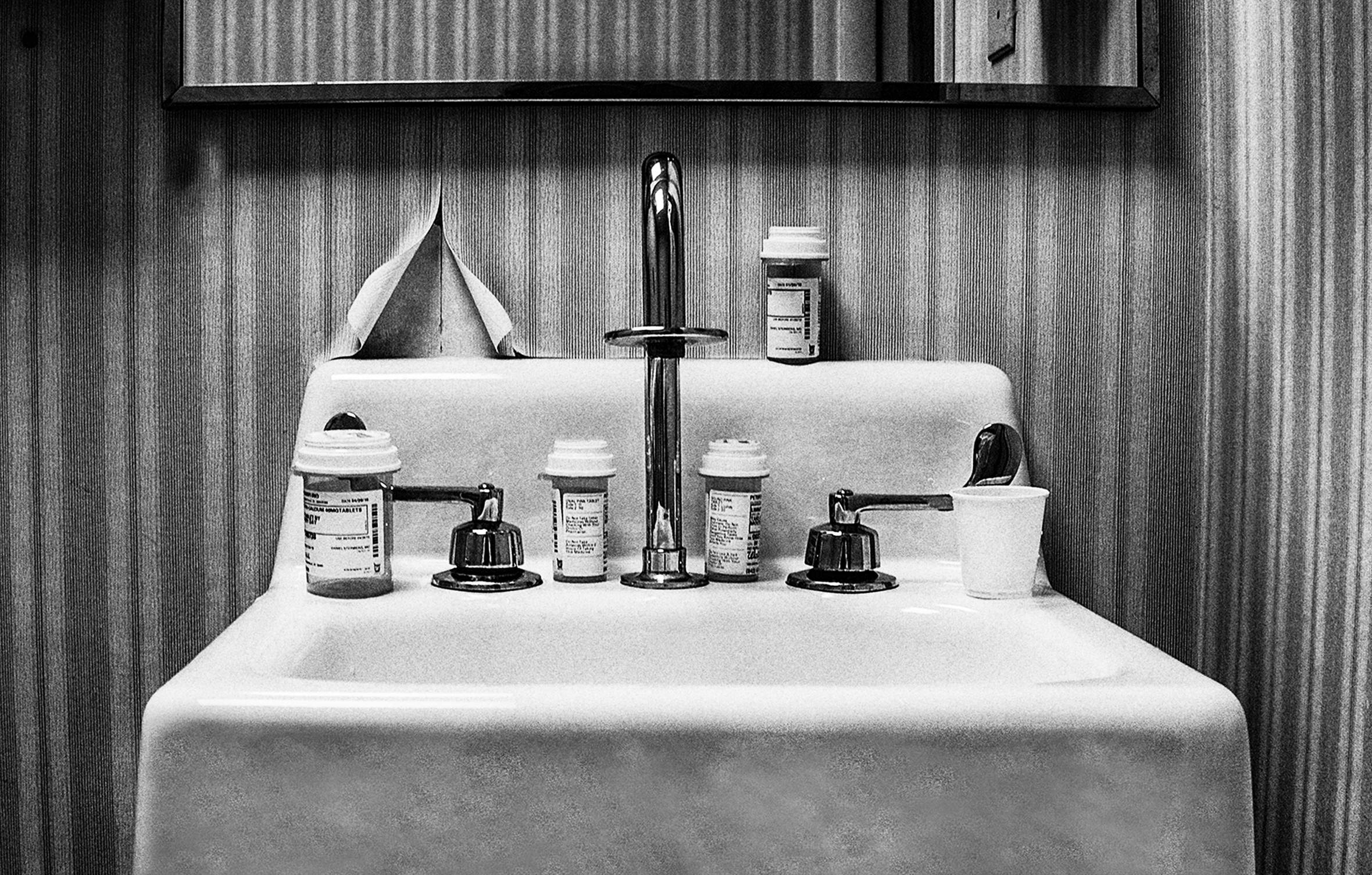 Close-up of the top of a small, white, pedestal bathroom sink with silver fixtures and the bottom part of a rectangular mirror against peeling, striped wallpaper. On the sink are pill bottles and a small plastic cup.