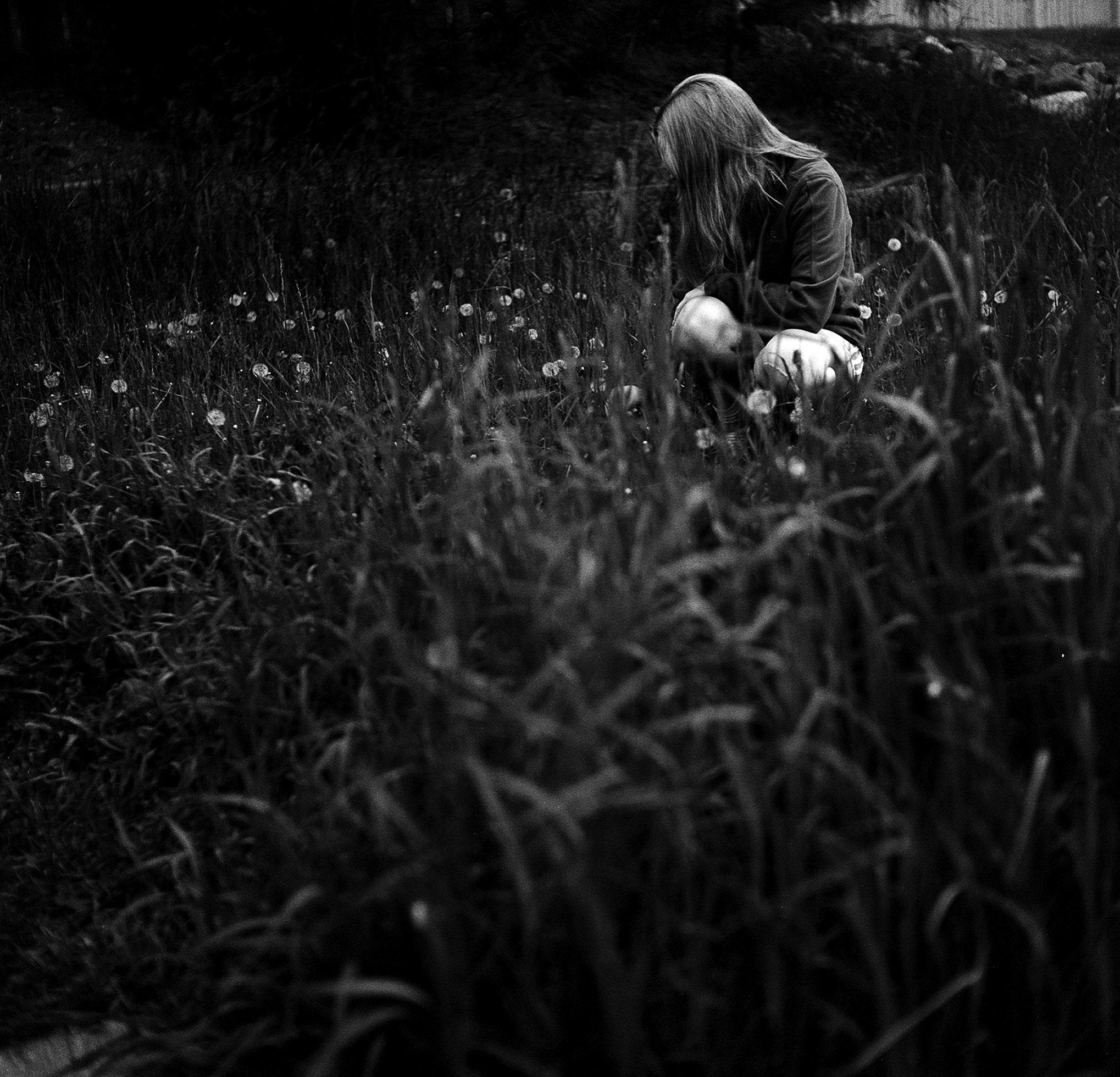 A woman dressed in shorts and a long-sleeve shirt crouches in a field of tall grass and wild flowers. She has long hair and looks to the side obscuring her face.