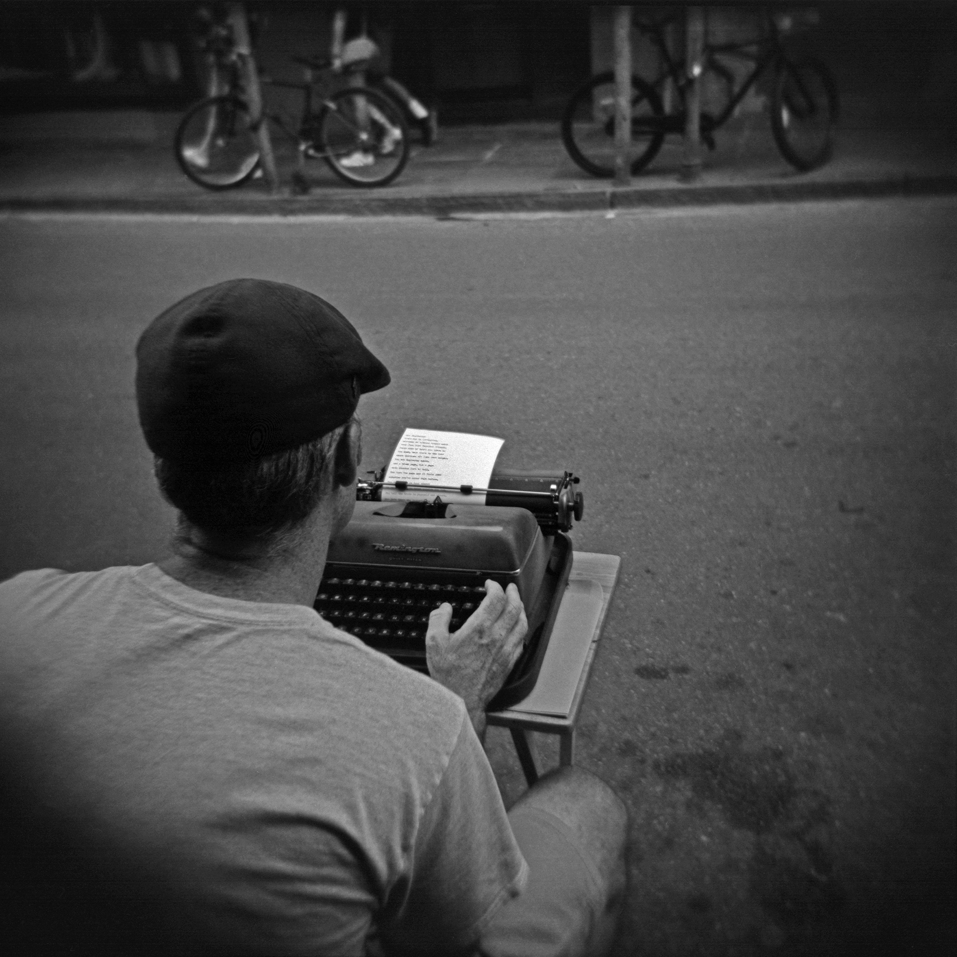 A man in a newsboy cap, T-shirt, and shorts sits at the side of a street typing on an old-fashioned typewriter on a small table. He is seen from the back. Across the street two bicycles are parked and people walk by on the sidewalk.