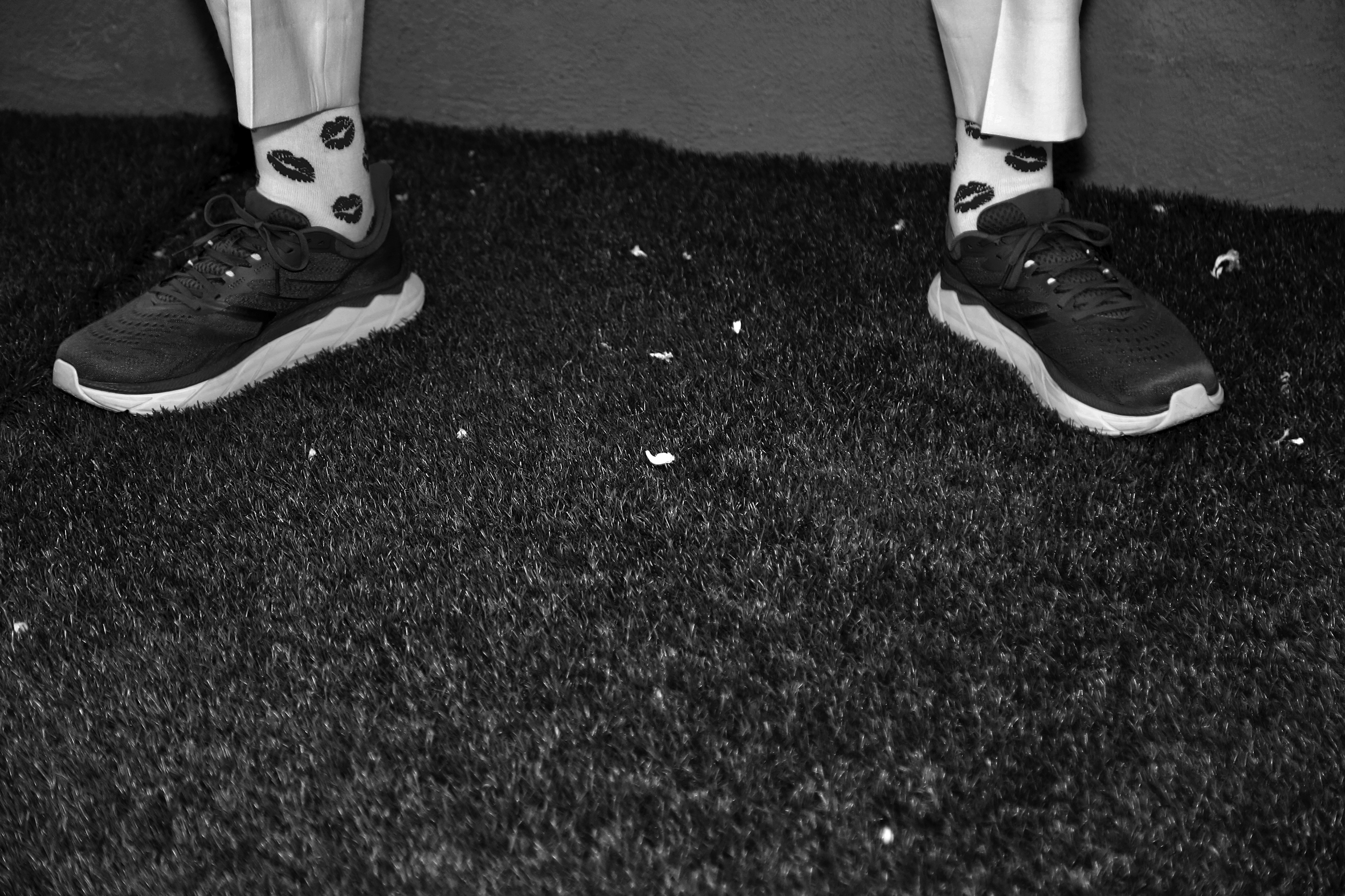 Close-up of a pair of men’s sneakers on the grass. The bottom part of his pants reach just above his ankles fully displaying white socks with a kissing-lips design.