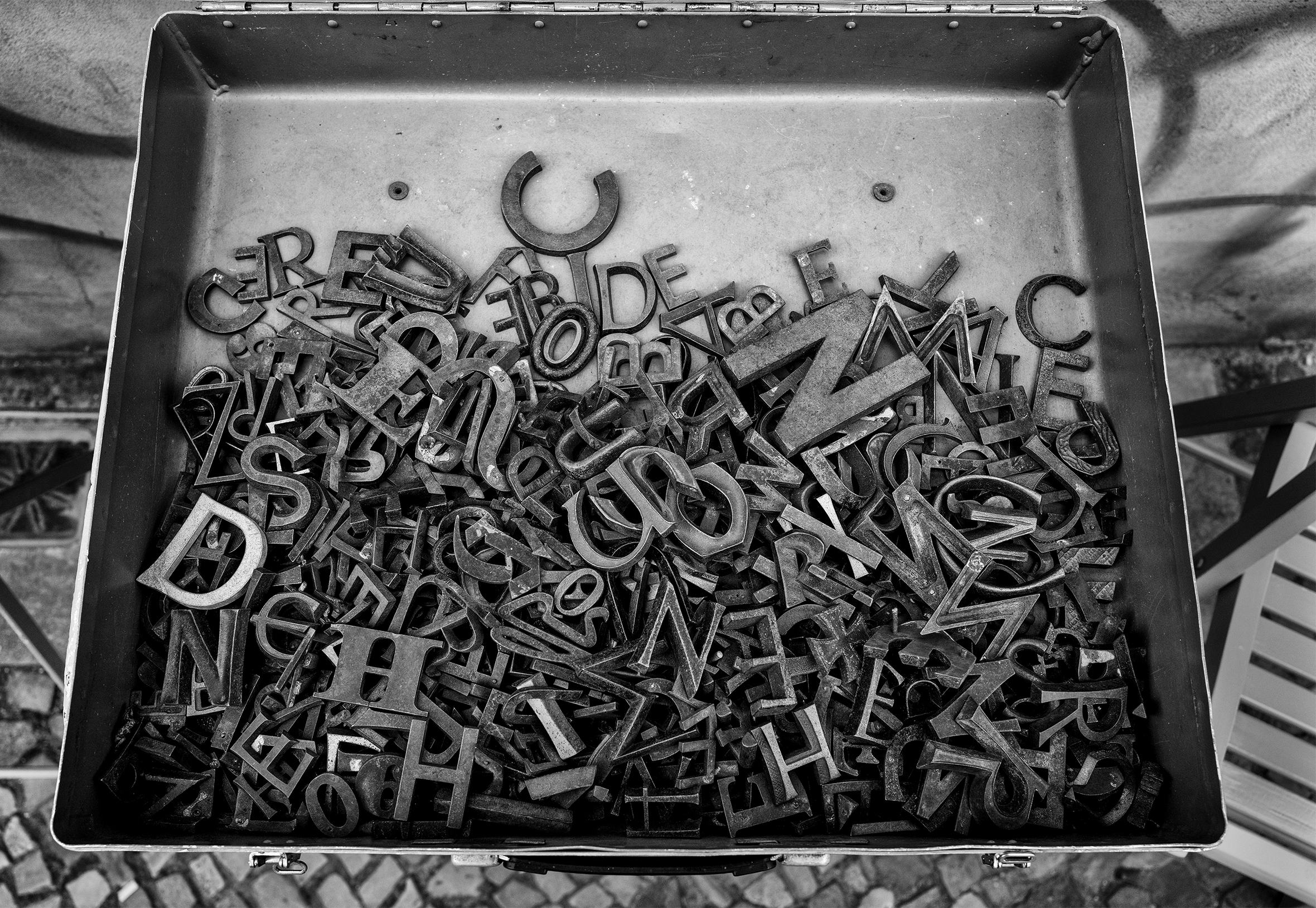 The bottom part of a hinged metal box half full of metal letters of various sizes.