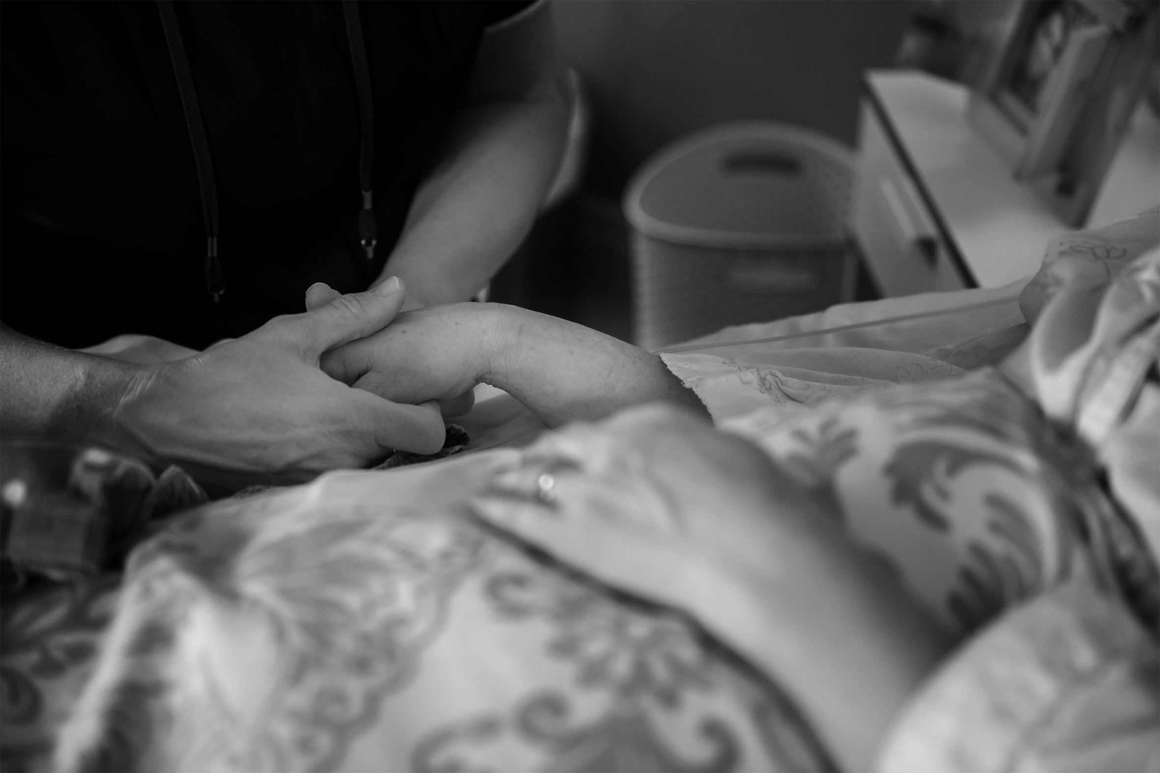 A person stands at a bedside and holds the right hand of the woman who is in bed. The focus of the image is on the hand-holding: the faces of the people are not visible.