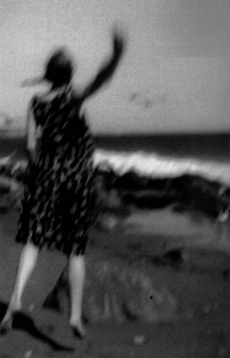 Blurry photo of a woman on the beach facing the water with her back to the camera. She is wearing a dress and is barefoot as she jumps up and waves her right hand with her arm fully extended.