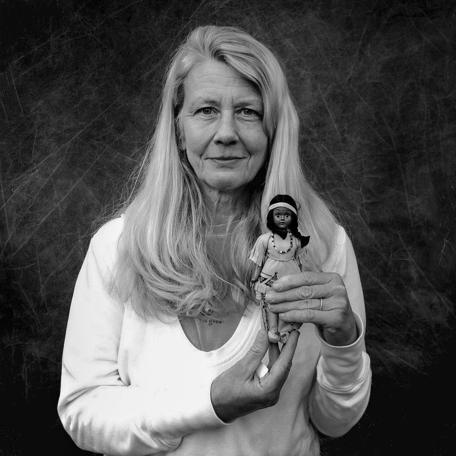 An older woman with a closed-mouth smile and long hair wearing layered T-shirts holds up a small Native American doll.