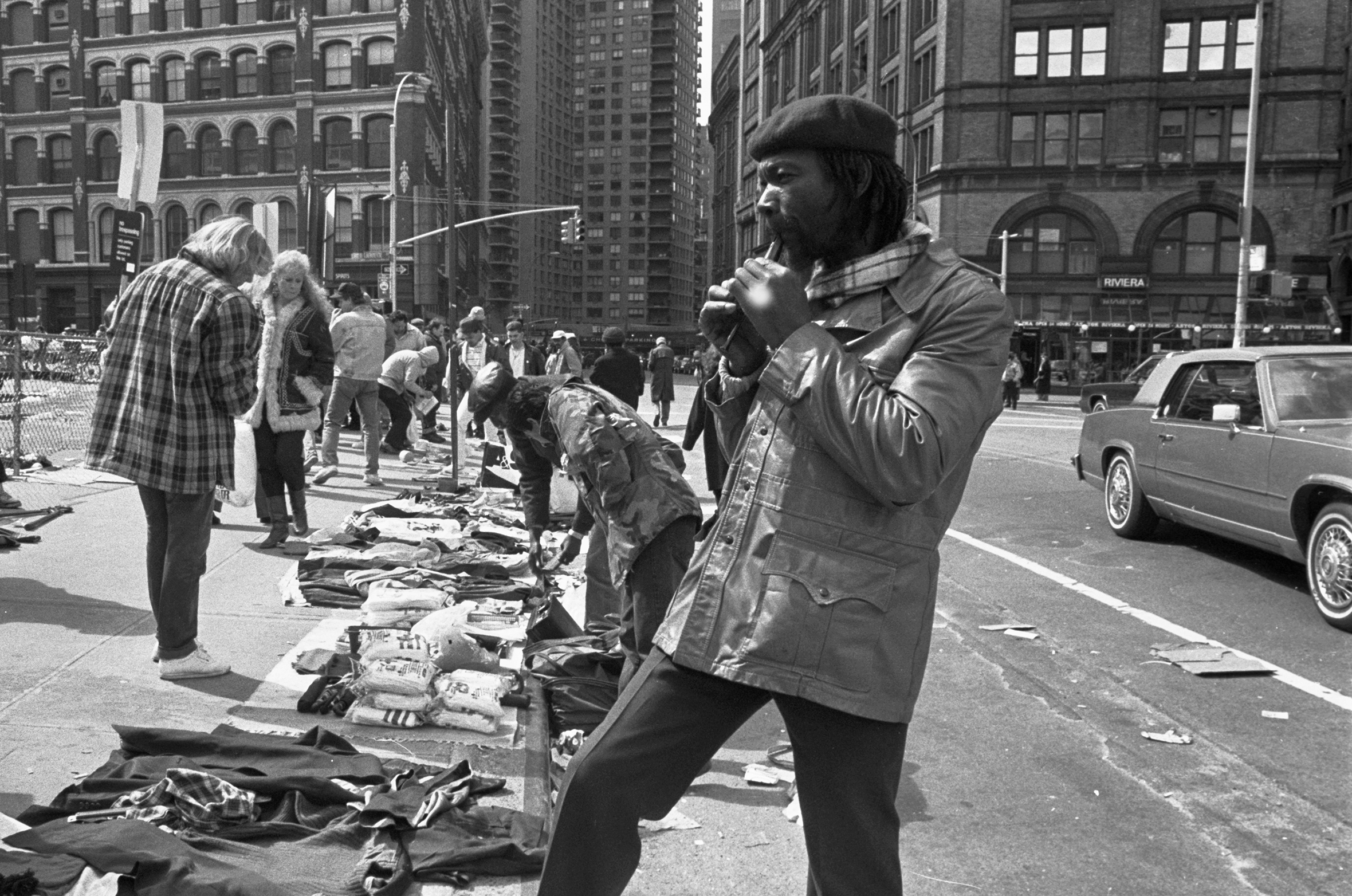 A sidewalk sale on a city street with many people looking at the merchandise actually down on the sidewalk. In the foreground a man with dreads wearing a beret and a leather coat plays what looks like a flutophone.