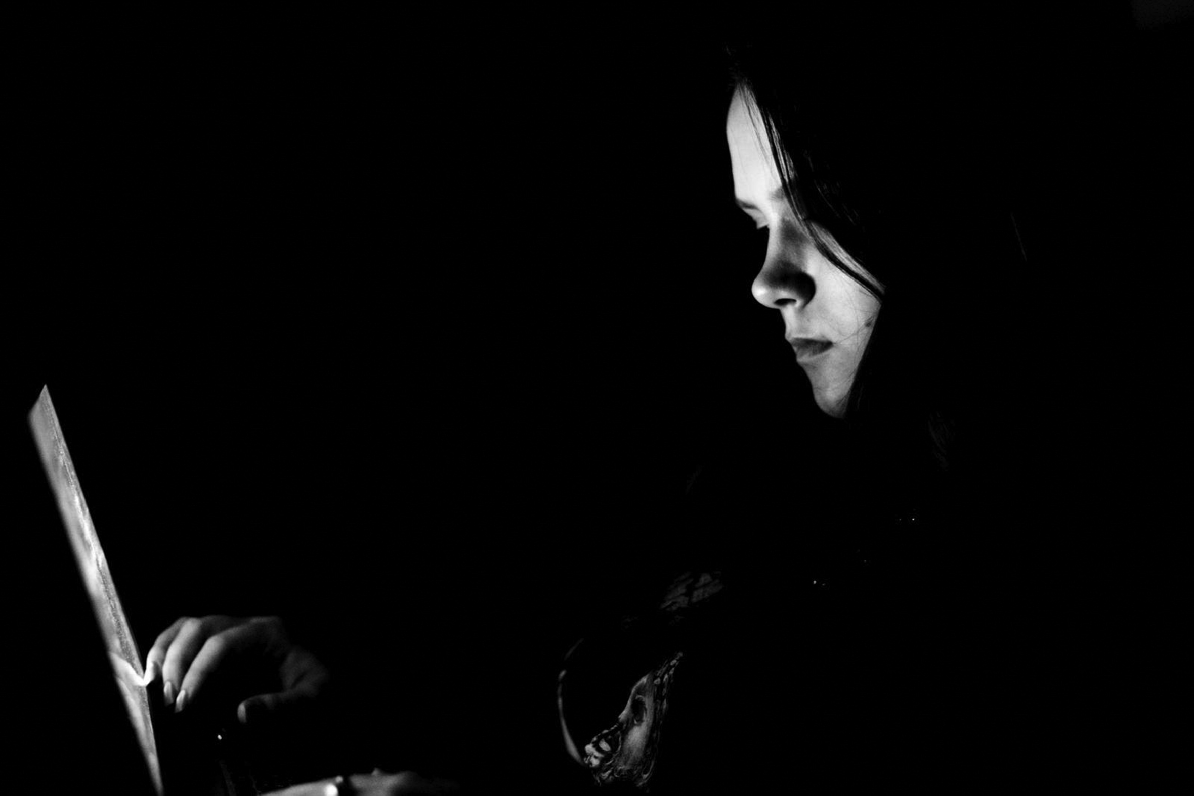 A young girl looks at her open laptop. The image is dark, but her face is illuminated by the computer screen.