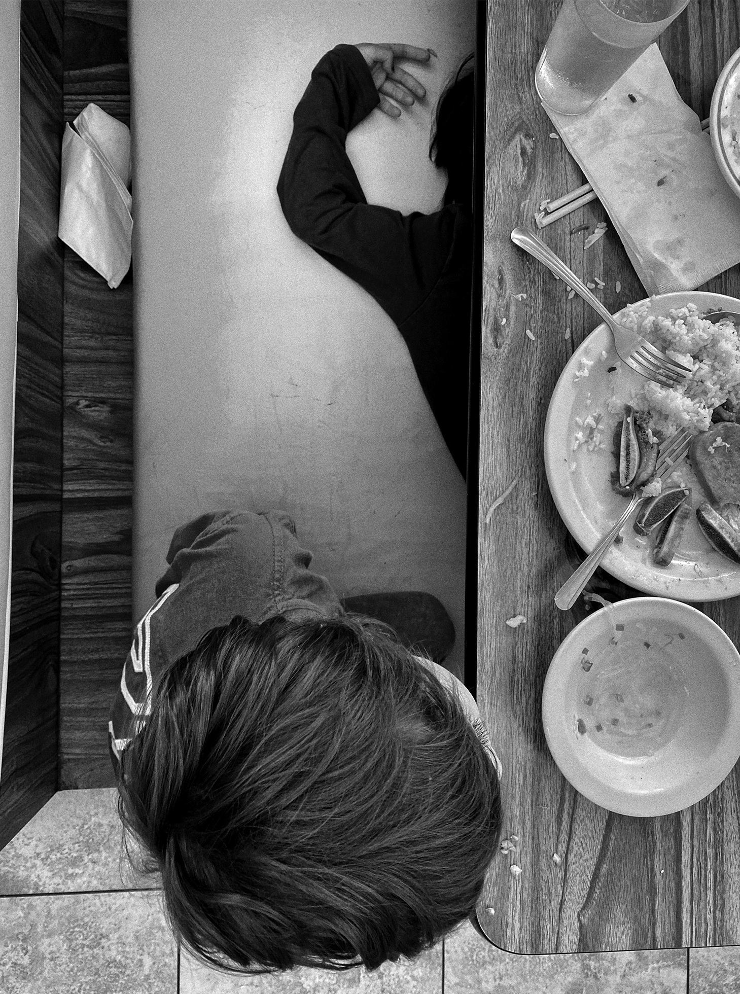 Above view of the head of a child sitting in a booth during a meal. To the left of the seated child is another child’s arm and part of their head and torso on top of the booth’s bench while the rest of them is obscured by the table.