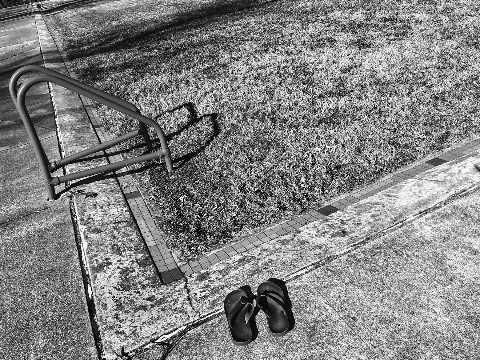 Close-up of an in-ground pool with a ladder that is completely filled in with grass growing on the top and a pair of flip-flops near the edge.