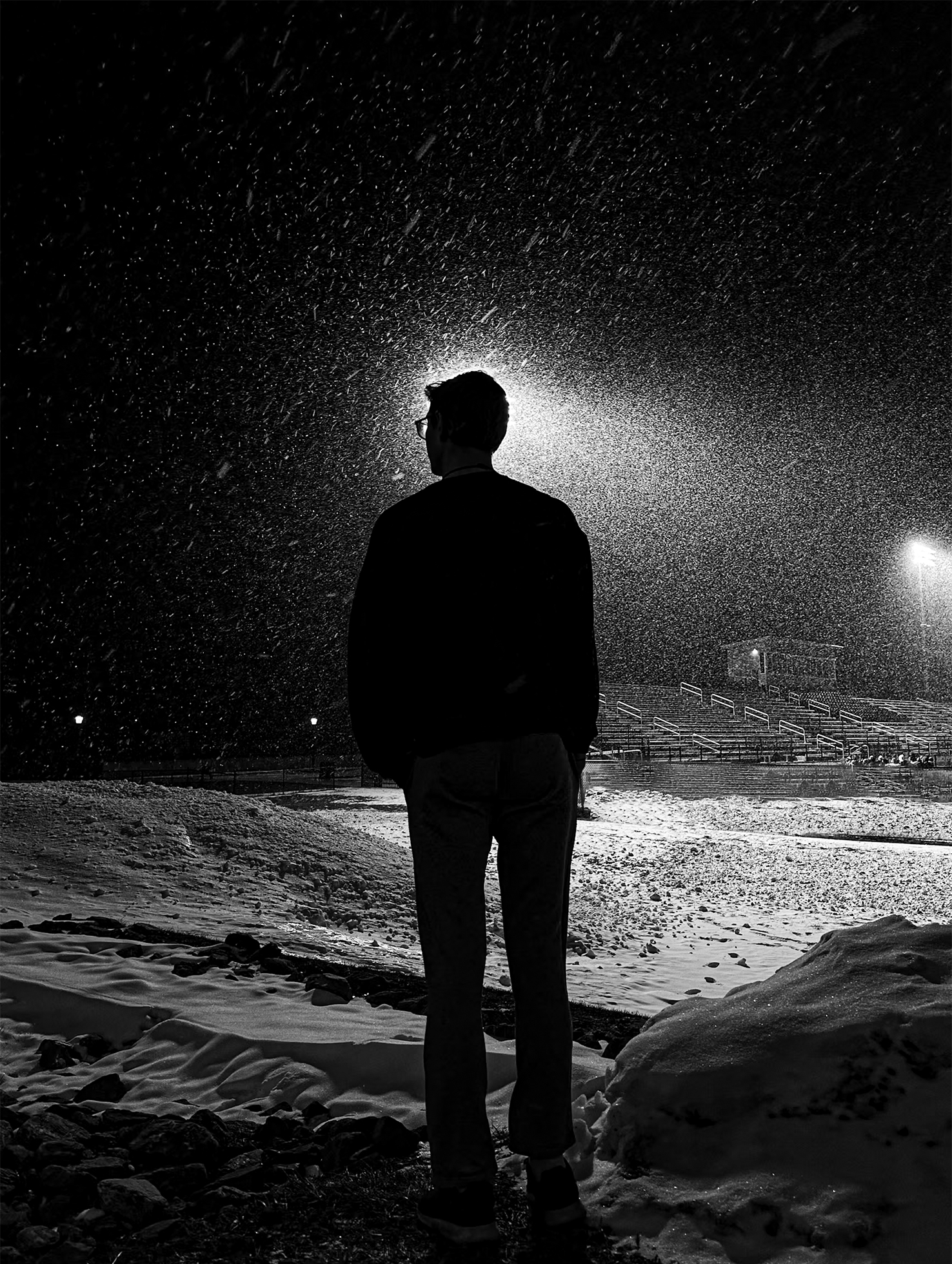 A man stands outside on a snowy night with his back to the camera near a field with bleachers. His head blocks the middle of one of the two distant floodlights such that there is a glow of light textured by the falling snow around his head.