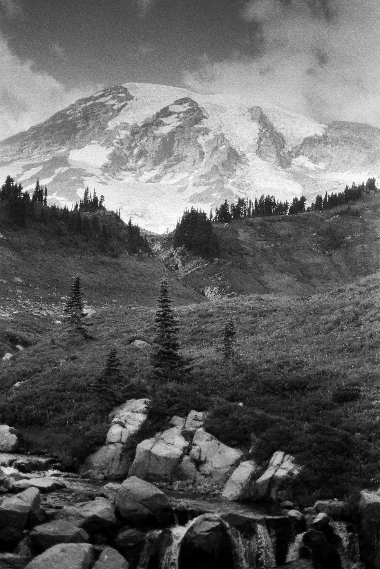 Boulders and hills with sparse evergreen trees in the foreground open to a snowy peak on a cloudy day.