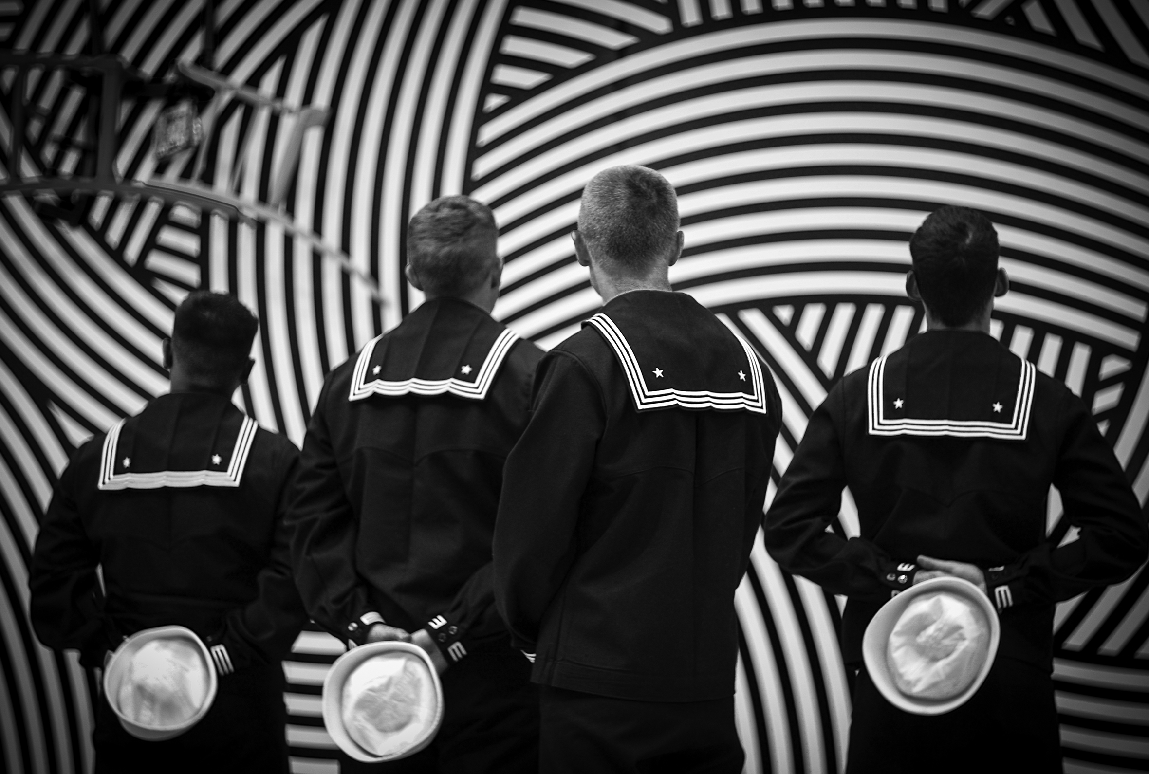 Four sailors in service dress blues seen from behind. Three have their hands behind their backs holding “Dixie cup” hats. They are in front of an art installation with curved, haphazard stripes and three interlocking chairs hanging from the top left.