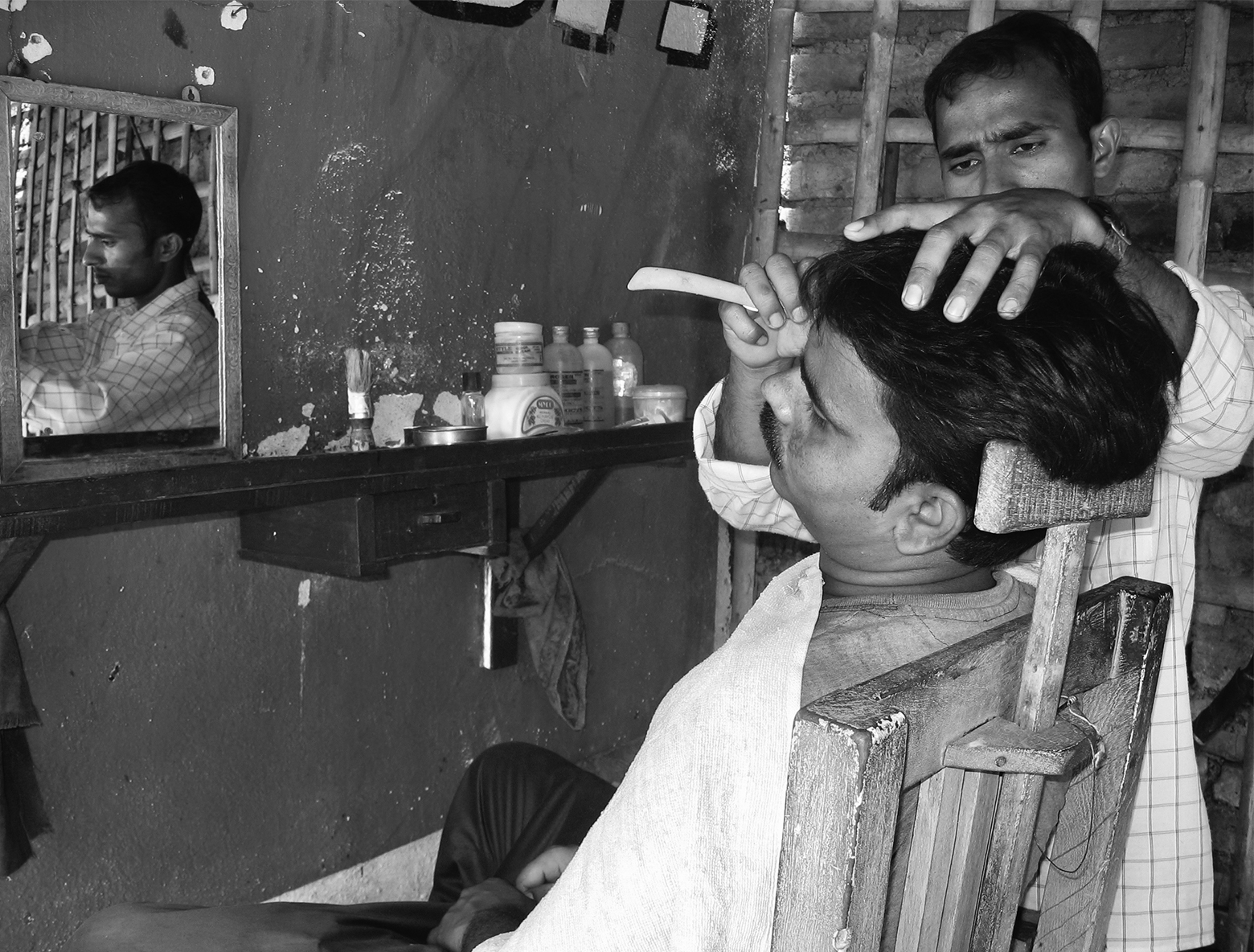 A barber shaves a man’s face with a straight razor while firmly grasping the top of the man’s head to steady it. The spare, wooden barber chair has a height-adjustable T-bar up the back against which the barber positions his client’s head.